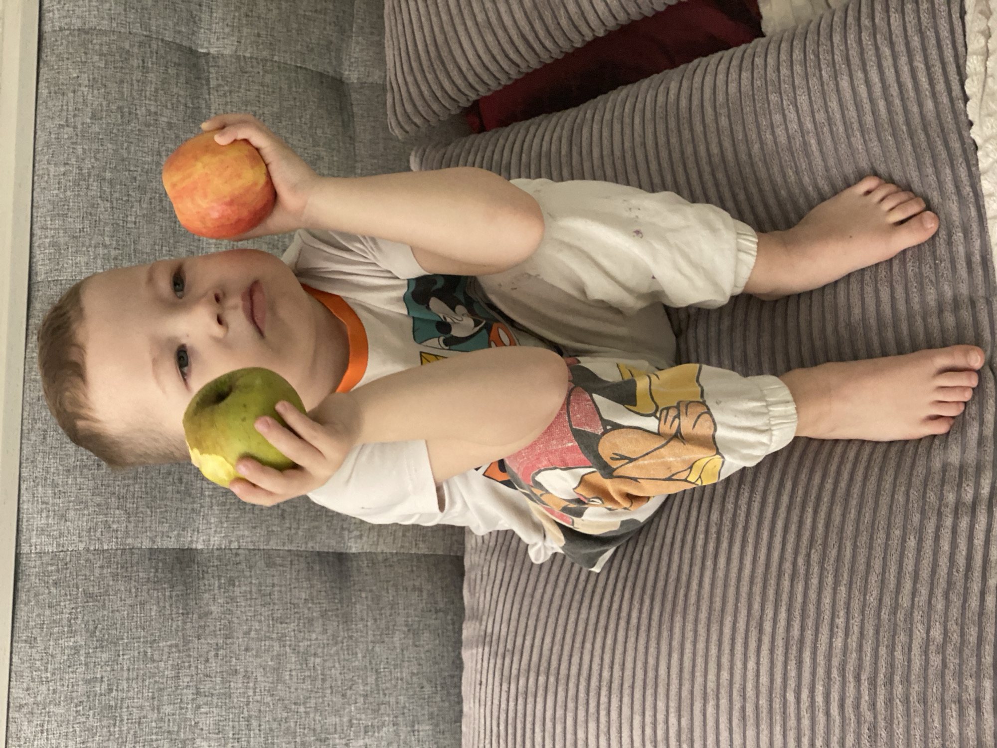 A child is reclining on a gray sofa, holding an apple in each hand. One apple is green, and the other is red and yellow. The child is wearing a white t-shirt with a cartoon character design and cartoon-themed pants. The sofa has a corduroy texture. The location is not discernible from the image.