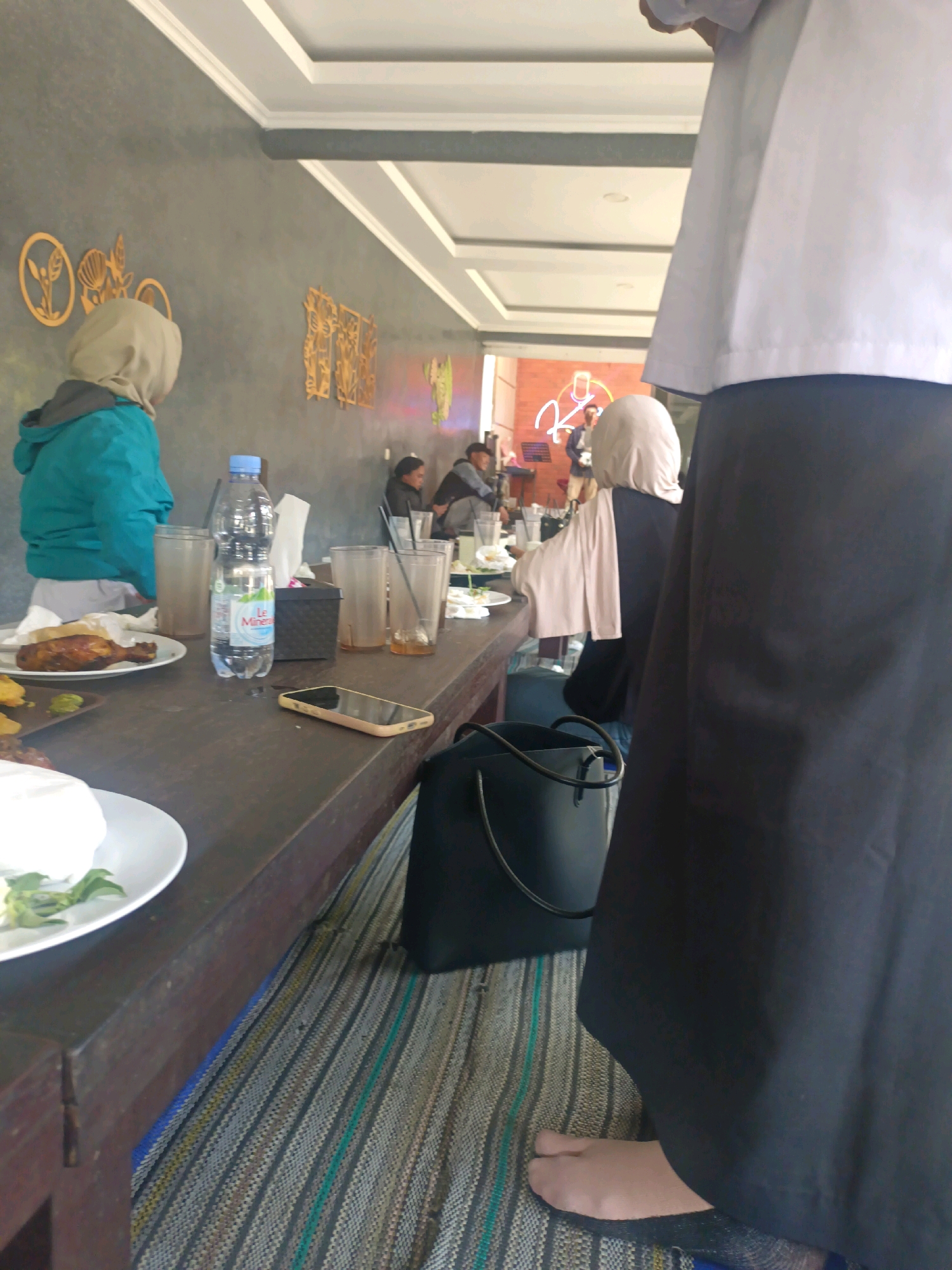 The image shows a long, dark wooden table set up in an indoor eating area, possibly a restaurant, in Kerangkulon, Indonesia. The table is covered with a matting material. On the table, there are plates of food, glasses, a bottle of mineral water, and a mobile phone. In the background, there is a wall with decorative panels. Several people are seated at the table, some wearing headscarves. One person in the foreground is partially visible, wearing a black garment and a white top. The scene suggests a casual dining or gathering setting.
