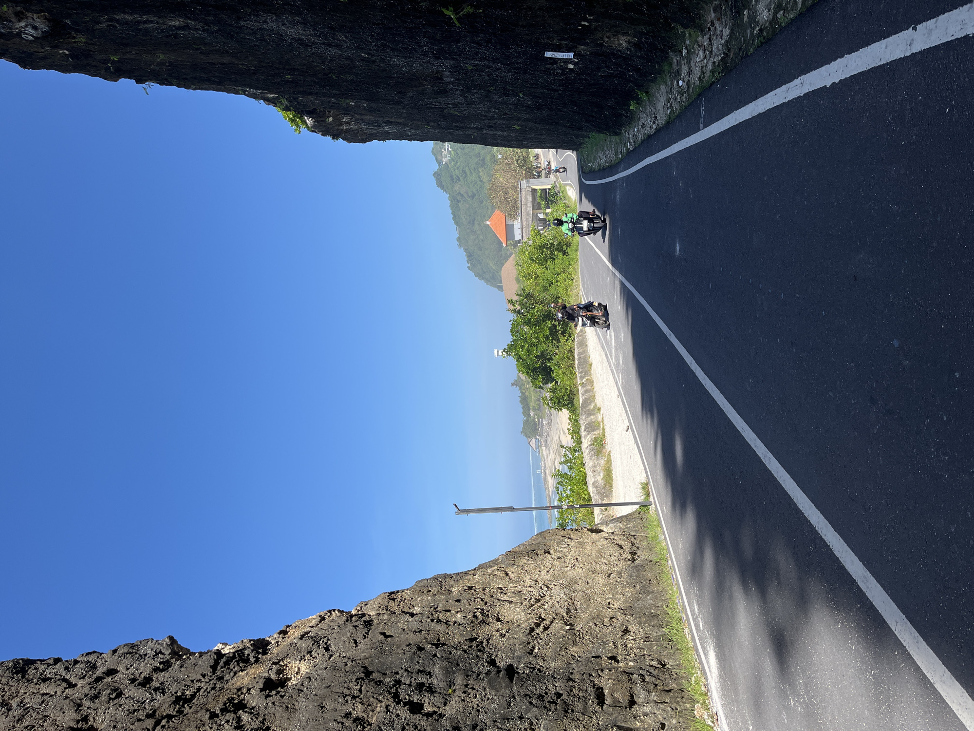 The image captures a road winding between rock cliffs in Kuta Selatan, Indonesia. The sky is clear and blue. The road has a white dividing line and shows shadows. Two motorcycles are visible on the road, one closer and one further away. In the distance, foliage and a building with an orange roof can be seen. To the left, a rugged rock cliff is visible, while to the right, a less imposing cliff stretches along the road. In the distance between the cliffs the ocean can be seen.