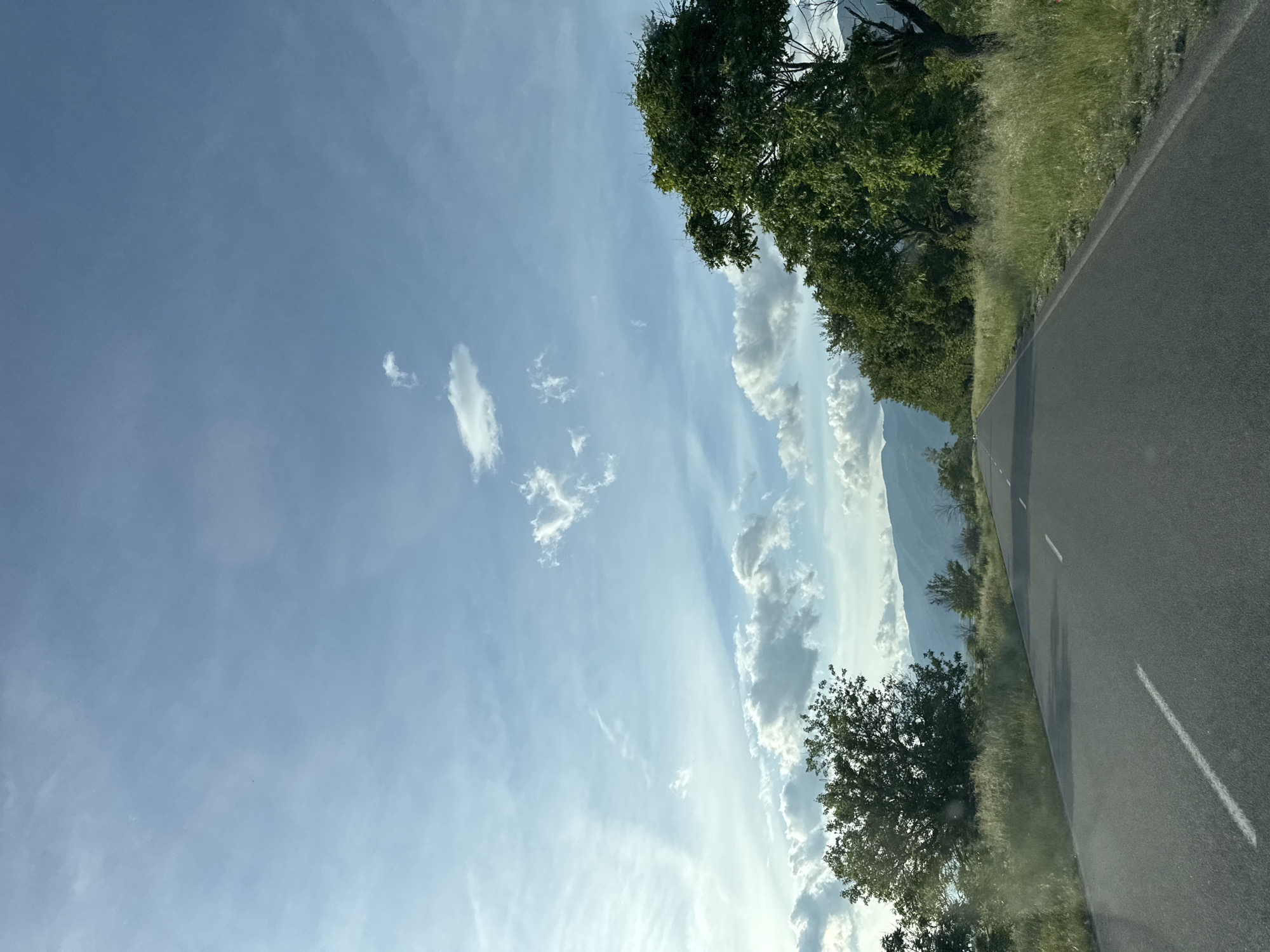The image shows a road under a partly cloudy sky, with trees lining the side of the road. In the distance, there is a mountain. The location is not discernible.