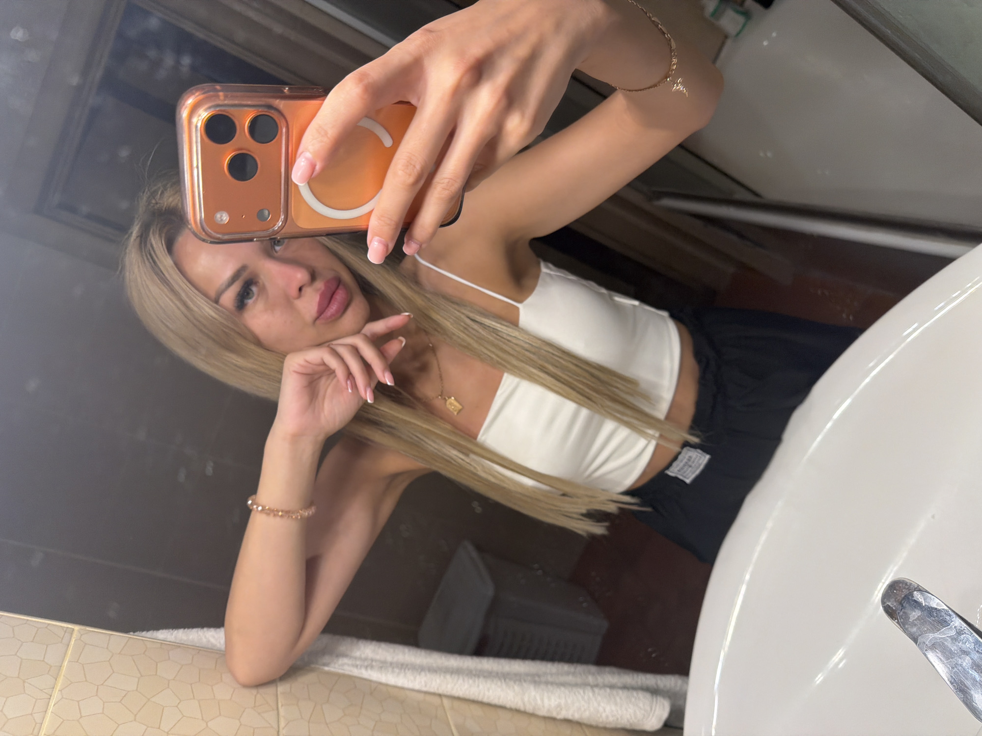 A young woman with long blonde hair takes a selfie in a bathroom. She is wearing a white crop top and black pants. She is holding her phone in her right hand, which has an orange case. Her left arm is resting on a tiled wall. The bathroom appears to be a home bathroom, with a sink in the foreground and a laundry basket and mirror visible in the background. The overall impression is a casual moment captured in a private setting. There are no specific indicators that place the location in Kyiv, Ukraine.