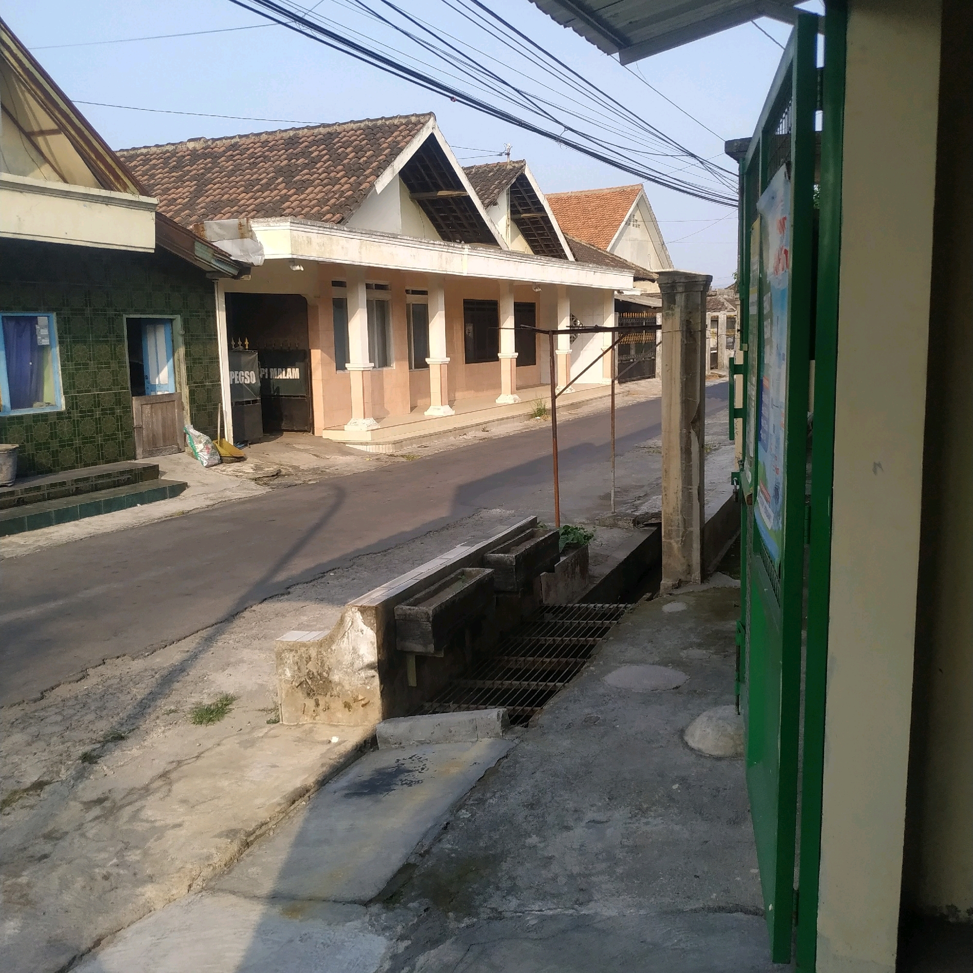 The image captures a street scene in Batan, Indonesia, on a clear day. In the foreground, a green metal gate is partially visible on the right, alongside a cream-colored wall. The street itself is paved with asphalt, with a concrete sidewalk running alongside it. To the left of the street, a drainage system with a metal grate is present. A concrete planter box, which appears to be part of the drainage system, holds a small amount of vegetation. Further down the street, several houses are visible, characterized by their tiled roofs and varying architectural styles. One house on the left has a dark green tiled facade, with a blue-tinted window and a wooden door. Adjacent to it, another building features a light peach-colored facade with white pillars. Electrical wires are strung across the sky overhead. In the background, more houses and buildings are discernible, suggesting a residential area within the city. The overall impression is that of a typical urban street in Indonesia.