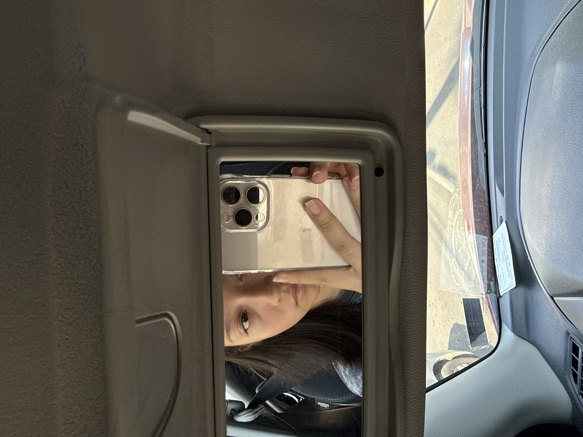 The image shows a selfie taken in the vanity mirror of a car sun visor. The reflected image includes the person taking the photo holding their phone, and their eye. The car's interior, specifically the dashboard and window are visible. A parking permit is affixed to the windshield. The precise location in Puebla, Mexico is not deducible from the image.