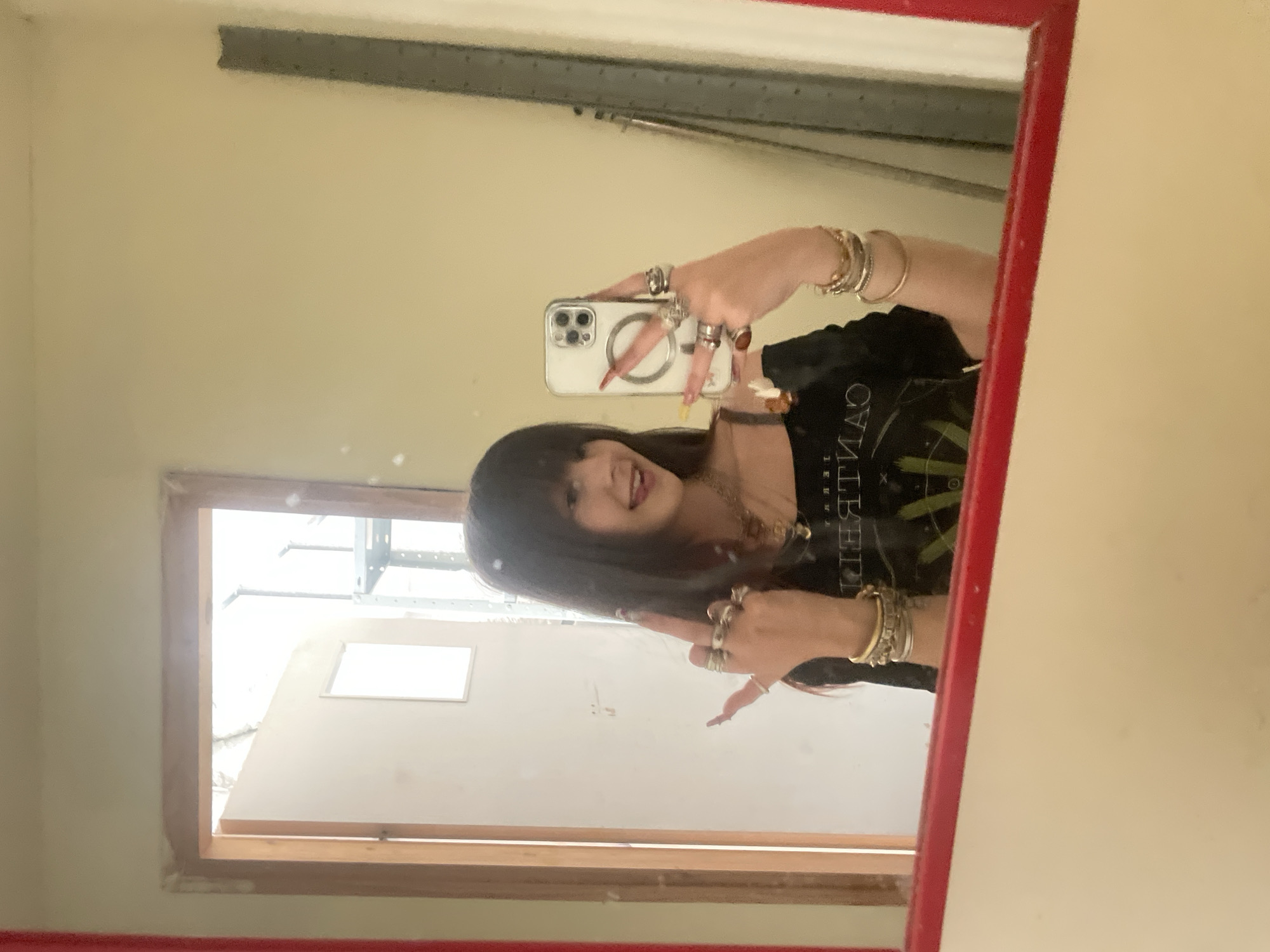 The image depicts a person taking a selfie in a mirror. The person has dark hair and is wearing a black t-shirt, multiple rings, and bracelets. The mirror has a red frame and reflects a room with a window, revealing a structure outside, possibly indicating an industrial or commercial area. The exact location in Raanana, Israel cannot be determined from the visual information.