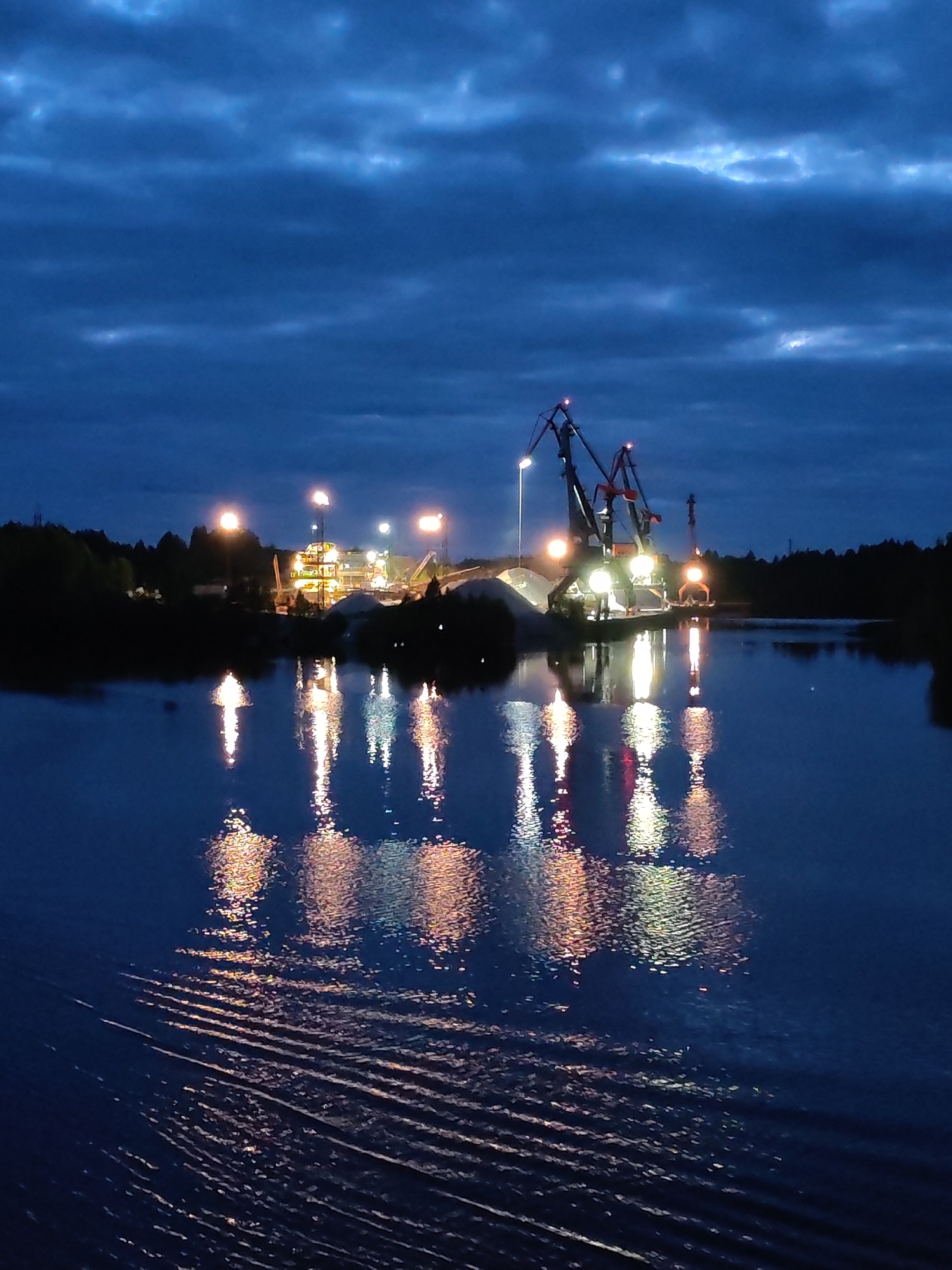 The image depicts a body of water at dusk or night, reflecting lights from a construction or industrial site on its far bank. The site features crane-like structures and brightly lit buildings, possibly a quarry or factory. The water surface exhibits ripples, indicating movement. On either side of the central illuminated area, darker silhouettes of trees suggest a wooded shoreline. This scene is located in Александровское, Russia.