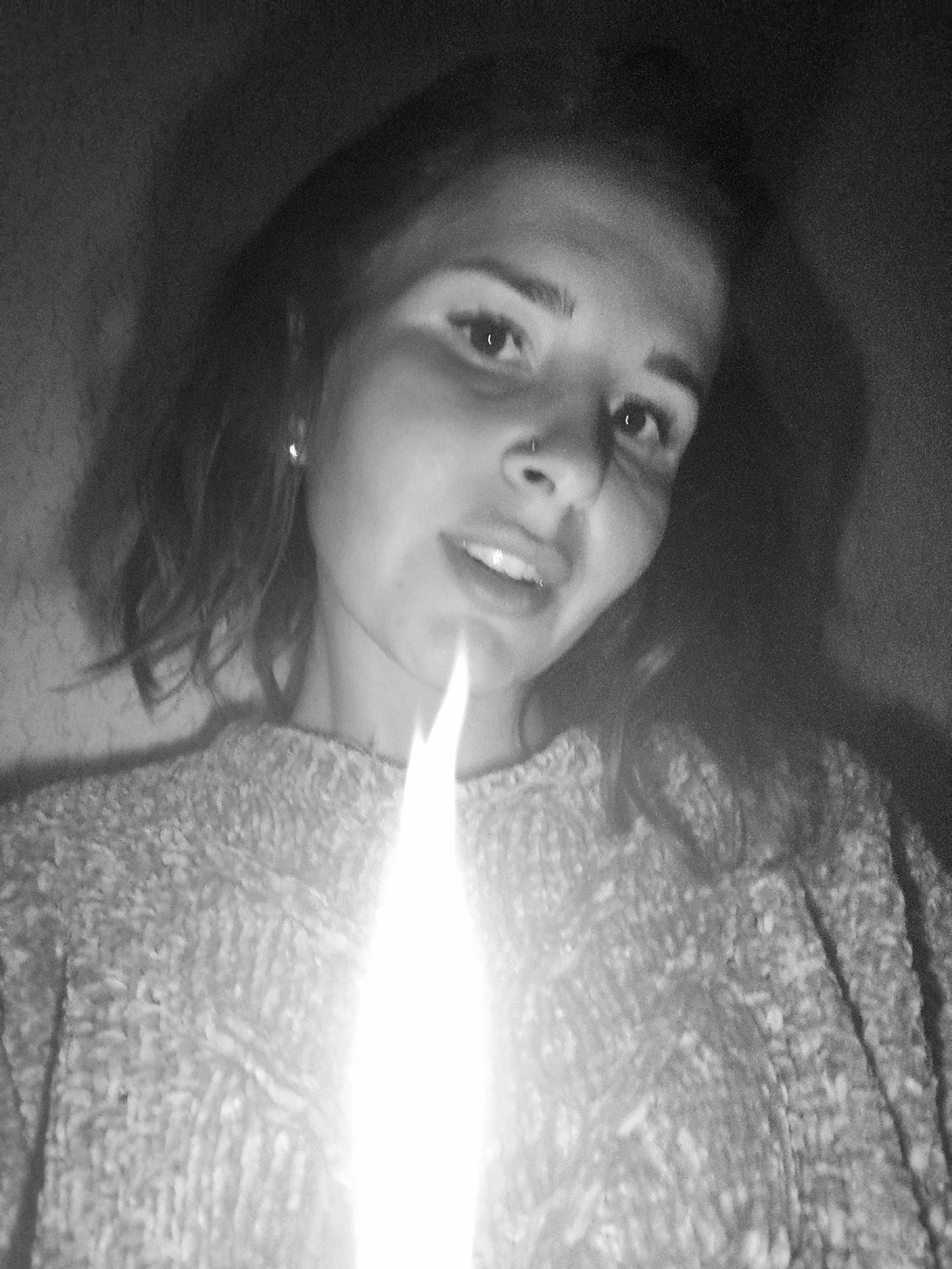 A grainy, monochrome, close-up portrait captures a young woman in an indoor, low-light setting, possibly at night, in Фоки, Russia. She is dramatically illuminated from below by a bright, vertical light source, resembling a candle flame, which extends upwards from the bottom edge of the frame to her chin.
The woman is looking directly at the viewer with a relaxed expression and a slight smile, her lips gently parted to reveal a hint of her teeth. She has medium-length, wavy hair that frames her face. Visible details include a small, reflective stud earring in her left ear and a silver nose ring in her right nostril.
She is wearing a textured, likely knitted or crocheted, sweater, the pattern of which is discernible across her shoulders and chest. The background is dark and largely indistinct, suggesting an unlit room, though a subtle texture on the wall behind her can be made out. The intense, solitary light source creates strong highlights on her face, reflecting in her eyes, and casts soft shadows, contributing to a moody and intimate atmosphere. There is no visible text in the image.