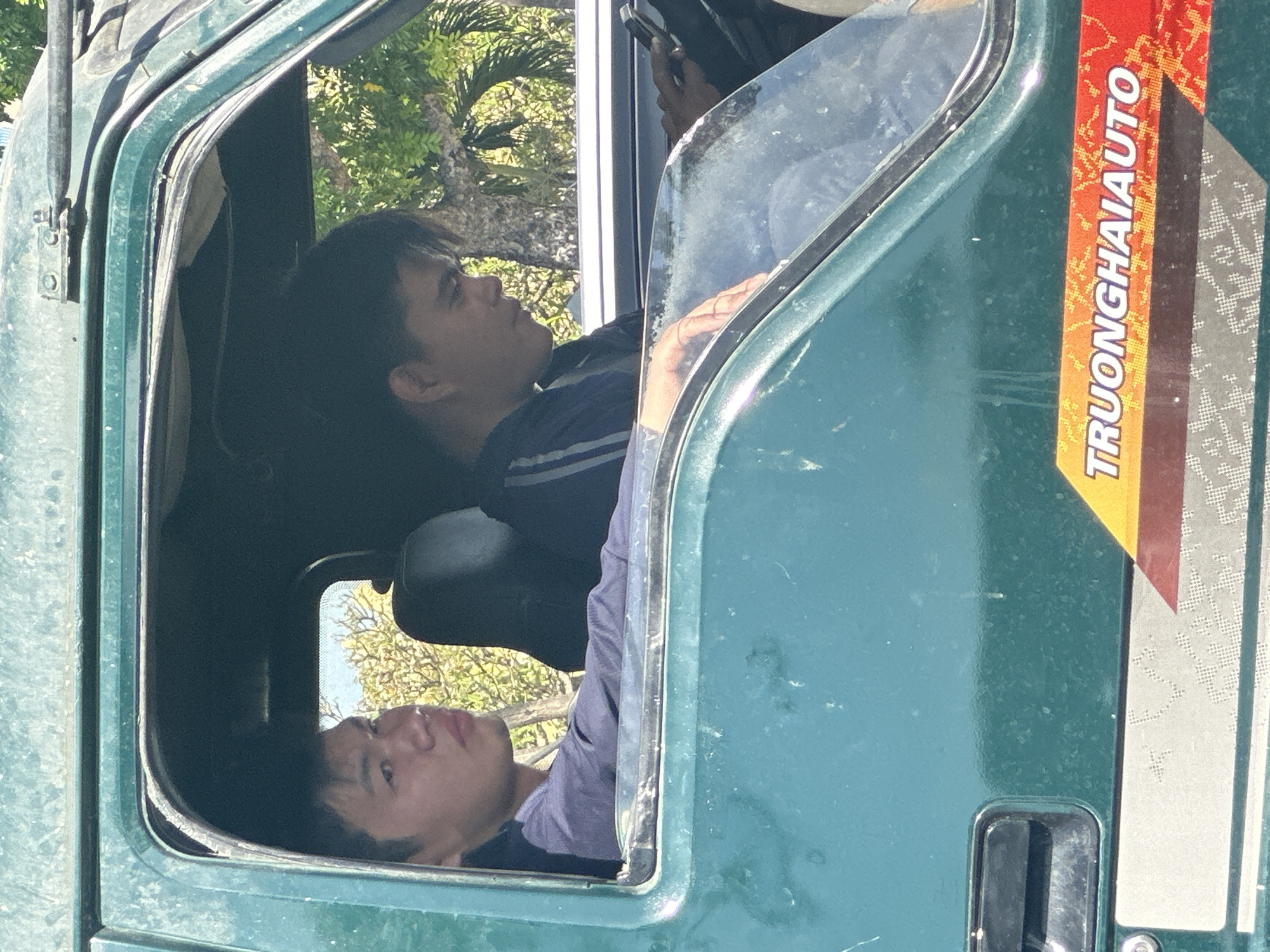 Two young males are seated inside the cab of a green utility vehicle. The individual on the left, with dark hair, is looking towards the camera with a neutral expression. The individual on the right, also with dark hair, is looking down and appears to be using a mobile phone. The vehicle's interior is visible, including dark-colored seats and a steering wheel. Outside the vehicle, the environment appears to be a bright, sunny day with trees and foliage visible in the background. A decal with the text "TRUONGHAIAUTO" is affixed to the side of the vehicle. The image was taken in a city setting, likely within Đà Nẵng, Vietnam, given the context of the decal. The overall scene suggests a moment of rest or transit for the occupants of the vehicle.