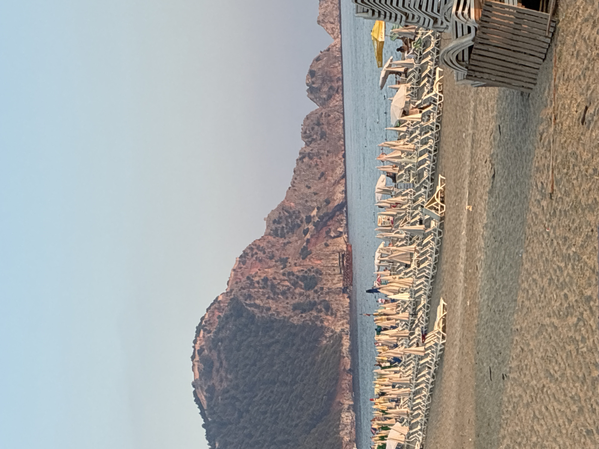 This is a vertical, eye-level medium shot of a busy beach scene in Alanya, Turkey. The image captures the expanse of a sandy beach lined with numerous rows of white lounge chairs, many of which are occupied by people. The chairs are arranged neatly, suggesting a well-organized beach resort.
Beyond the chairs, the calm, blue sea stretches out to meet the horizon. In the distance, imposing, rocky mountains rise from the landscape, their rugged textures and varied shades of brown and gray dominating the background. The sky above is a clear, pale blue, indicating fair weather, likely late afternoon or early evening given the warm, soft lighting that casts subtle shadows.
The overall mood is one of relaxation and leisure. The presence of many lounge chairs and the calm sea suggest a popular tourist destination. There are no visible signs of text in this image. The orientation of the image is vertical, giving a sense of height and depth to the scene.