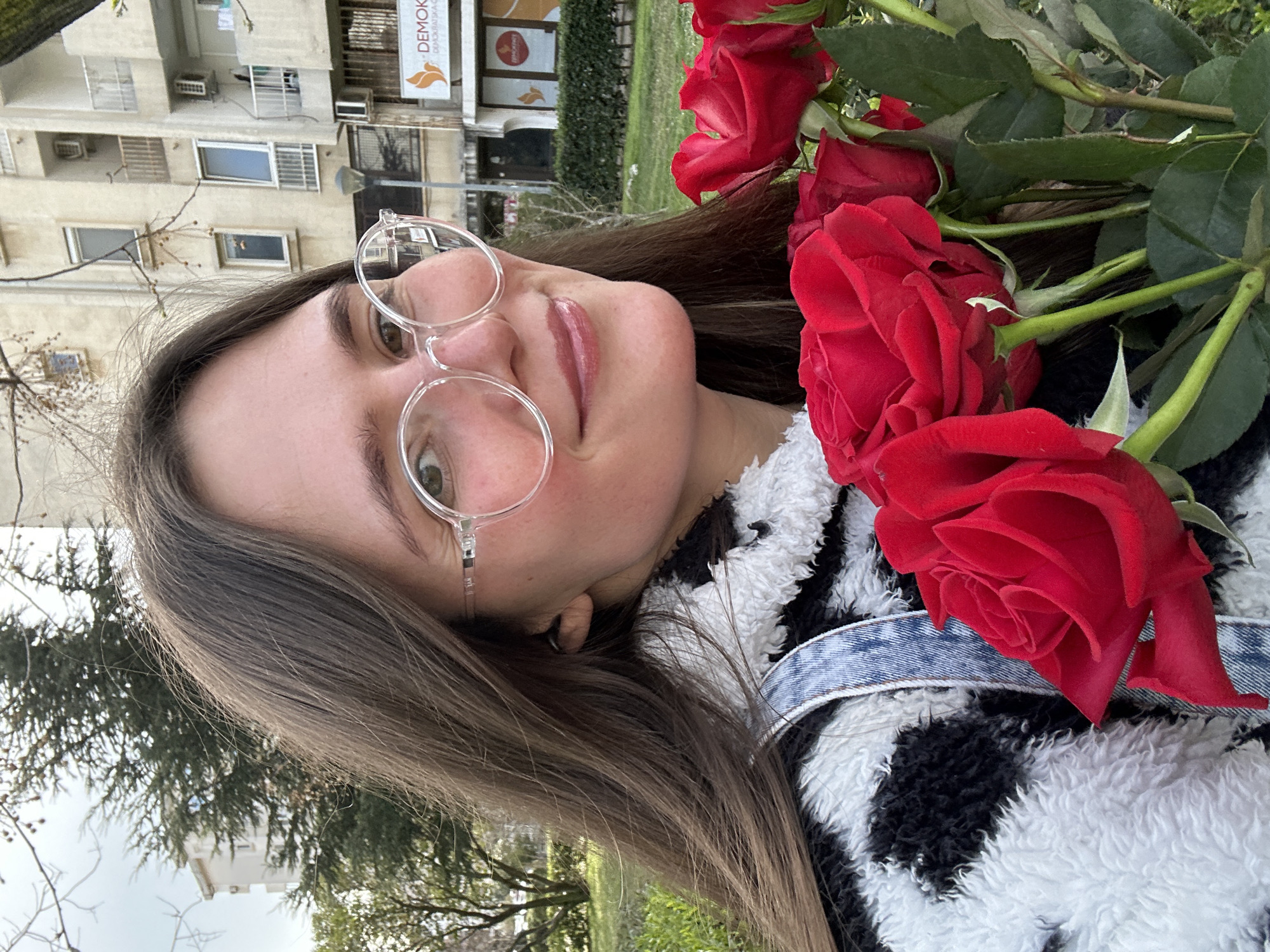 A young female subject is depicted in a close-up, outdoors, within the city of Šušanj, Montenegro. She has long, straight, dark brown hair, parted to her left, and wears clear-framed circular spectacles. Her facial expression is a gentle smile, with visible lips. She is dressed in a black and white patterned, shaggy fleece garment, and a blue denim strap is visible over her right shoulder. The subject holds a bouquet of approximately ten red roses, prominent in the lower right and center of the frame, featuring red petals and green stems with leaves.
The background consists of an urban environment under an overcast sky. To the subject's left, a light-colored multi-story building is partially visible through the bare branches of deciduous trees and green coniferous trees. To her right, a light-colored multi-story residential building with balconies and several exterior air conditioning units is present. Below this building, a green hedge is visible. A lamppost stands in the middle ground. Further right, a commercial building features a sign with an orange bird/leaf logo and partially visible text "DEMOKRATSKA CRNA GORA," identifying a Montenegrin political party.