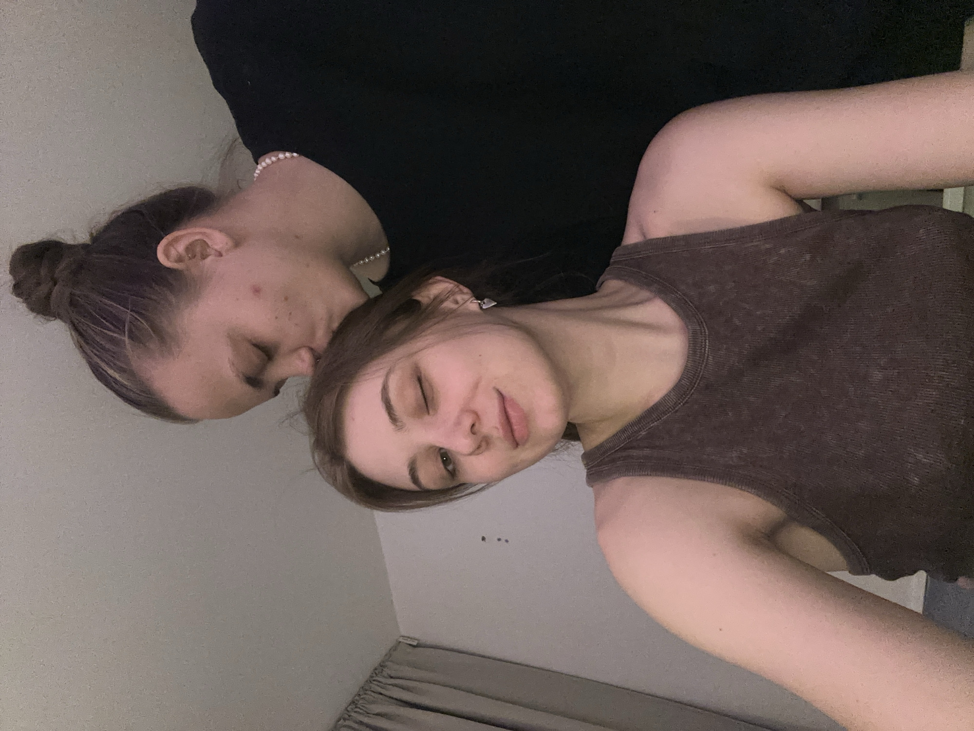 Two young women are posing indoors. One woman has brown hair in a bun and is wearing a black top and a pearl necklace. She is close to the other woman, possibly kissing her head. The other woman has brown hair, one eye closed in a wink, and is wearing a brown tank top. The background includes a plain wall and a glimpse of a curtain. There is no evidence of location in the city of Moscow, Russia.