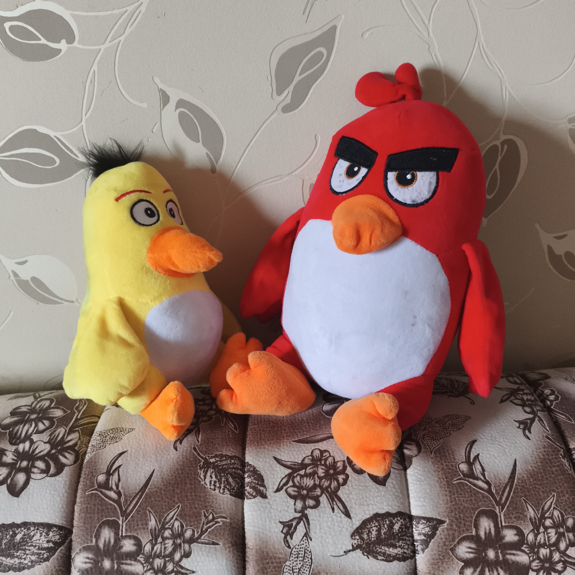 Two plush toys, depicting characters from the Angry Birds franchise, are seated side-by-side on a patterned sofa, leaning against a wallpapered wall. The indoor setting is likely a living room or bedroom in Chaikovsky, Russia.
On the left is a yellow bird plush toy, resembling Chuck. It has black, spiky hair on its head, large white eyes with black pupils, and prominent red eyebrows that give it a slightly surprised or curious expression. It features an orange beak, small yellow wings, and orange feet.
To its right sits a larger red plush toy, clearly identifiable as Red. This toy displays Red's characteristic angry or determined expression, conveyed by thick, downward-sloping black eyebrows over its large white eyes, which have visible brown irises and black pupils. It also has an orange beak, small red wings, and orange feet.
The sofa they rest upon is upholstered with a fabric featuring a repeating floral pattern in shades of brown, beige, and white. The wall behind the sofa is covered in textured wallpaper with a light beige-grey background, adorned with subtle, lighter grey or white abstract leaf and vine patterns. The lighting is bright and even, suggesting a daytime setting indoors. No specific activity is occurring beyond the toys being posed for the photograph. No text is visible in the image.