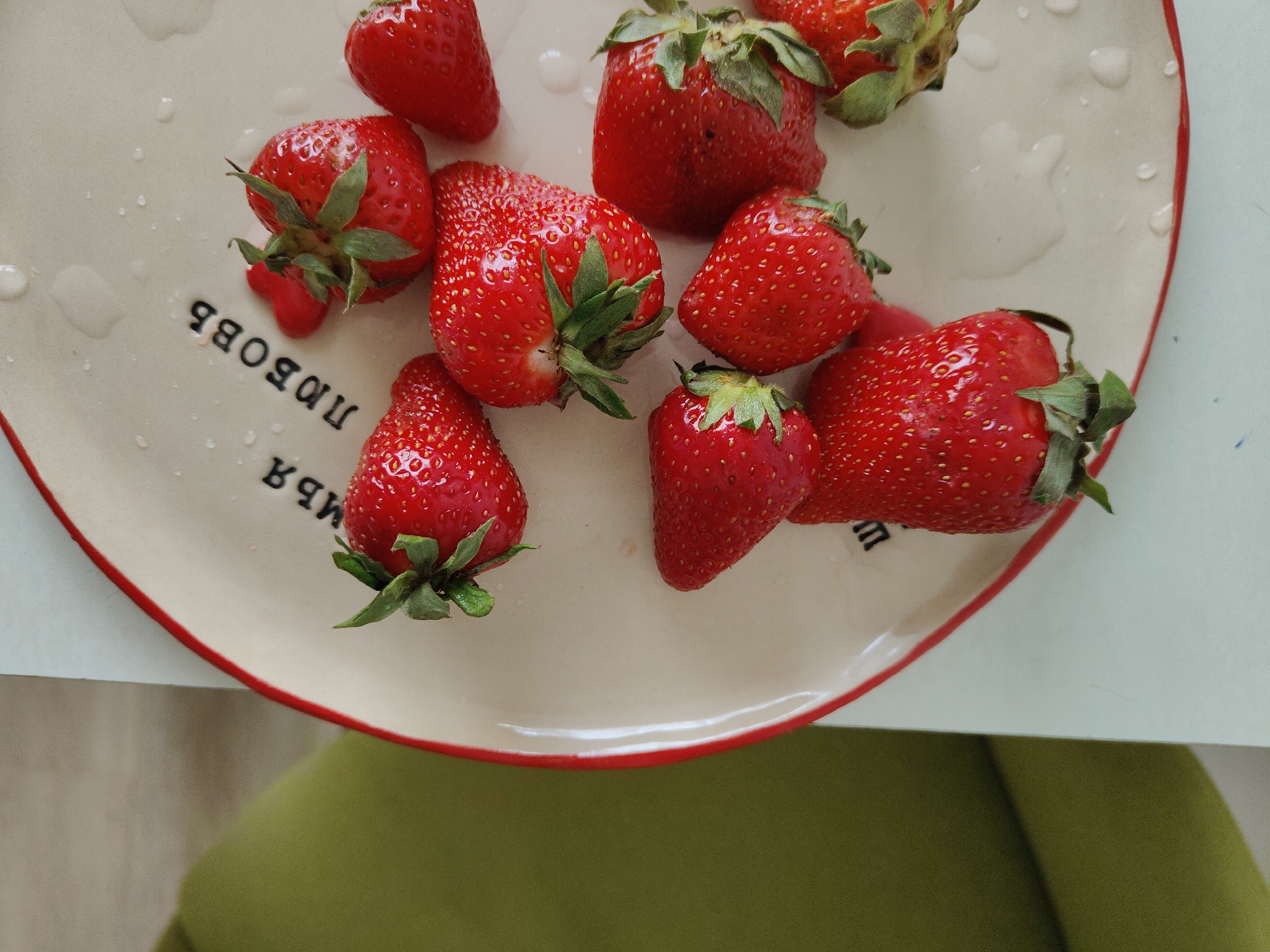 A close-up shot shows a plate of strawberries on a green surface. The plate is cream-colored with a red rim. It has the word "ЛЮБОВЬ" written on it. Seven strawberries are visible, arranged haphazardly on the plate. Some of them have water droplets on them. The background is out of focus.