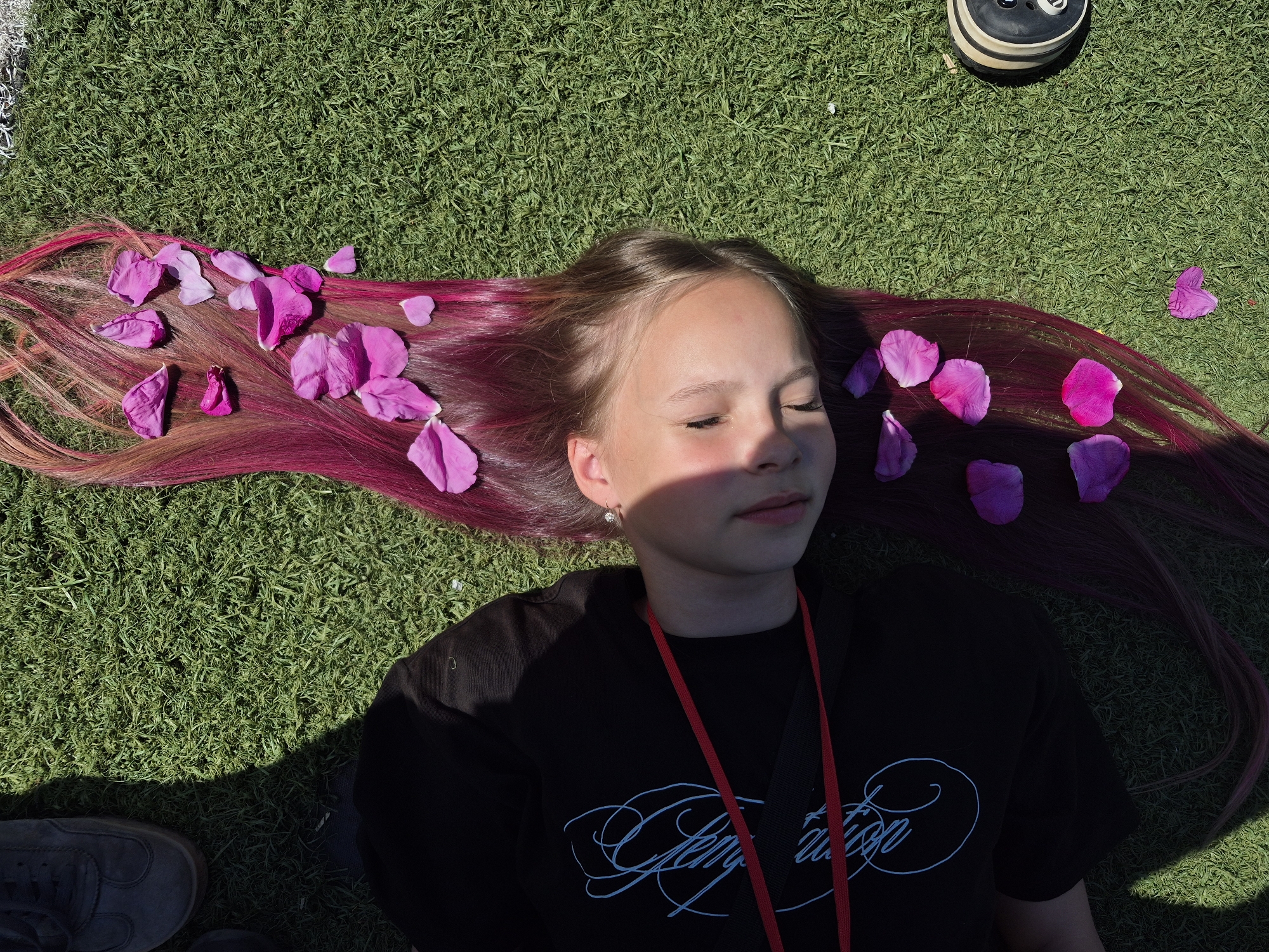 A young girl lies on her back on a bright green, artificial turf surface, her eyes closed. Her long, dyed hair, a mix of blonde and vibrant pink, is spread out beside her, adorned with scattered pink and purple flower petals. She wears a black t-shirt with a light blue graphic design and a red strap is visible across her chest. Sunlight casts a shadow across her face and torso. To her left, a person's legs are visible, wearing dark sneakers. To her right, the edge of a black and white shoe is visible. The scene appears to be outdoors, possibly in a park or recreational area in Chaikovsky, Russia, given the artificial turf.