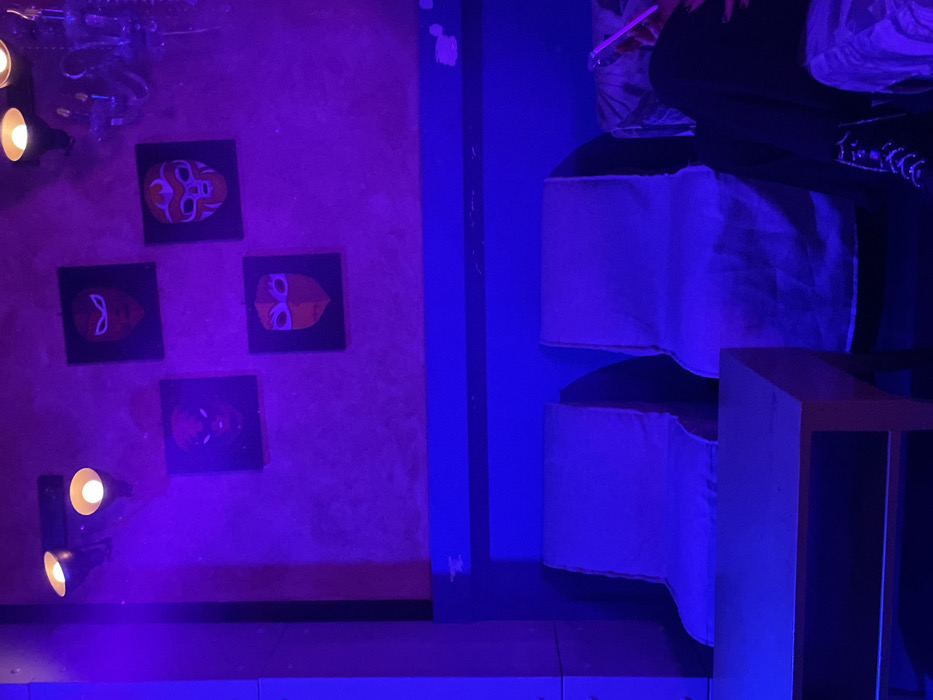 The image captures an indoor scene, likely a lounge or bar, characterized by strong purple and blue ambient lighting. The setting creates a vibrant, nocturnal atmosphere in Drapetsona, Greece.
On the left, a textured wall, possibly concrete, is illuminated with a deep purple hue. Adorning the wall are four square artworks arranged in a diamond pattern, each depicting a stylized, colorful mask or face, possibly resembling Lucha Libre masks with orange, red, and white details against a dark background. Below these artworks, two sets of double-barrel spotlights are mounted, emitting warm, yellow light upwards, contrasting with the cool ambient lighting.
Separating the wall from the right side of the scene is a vertical section, possibly a column or partition, bathed in a deep blue light. To the right, a person is seated on a stack of light-colored, possibly white or off-white, cylindrical cushions or poufs. The person's lower body is visible, wearing dark trousers and dark shoes with some white detailing. They are holding a glowing, translucent pink/purple stick-like object, suggesting a light stick or an illuminated drink accessory.
In the foreground, a light-colored, possibly pale blue or grey, raised platform or floor section is visible. The overall impression is one of a contemporary and themed entertainment venue. There is no visible text in the image.
