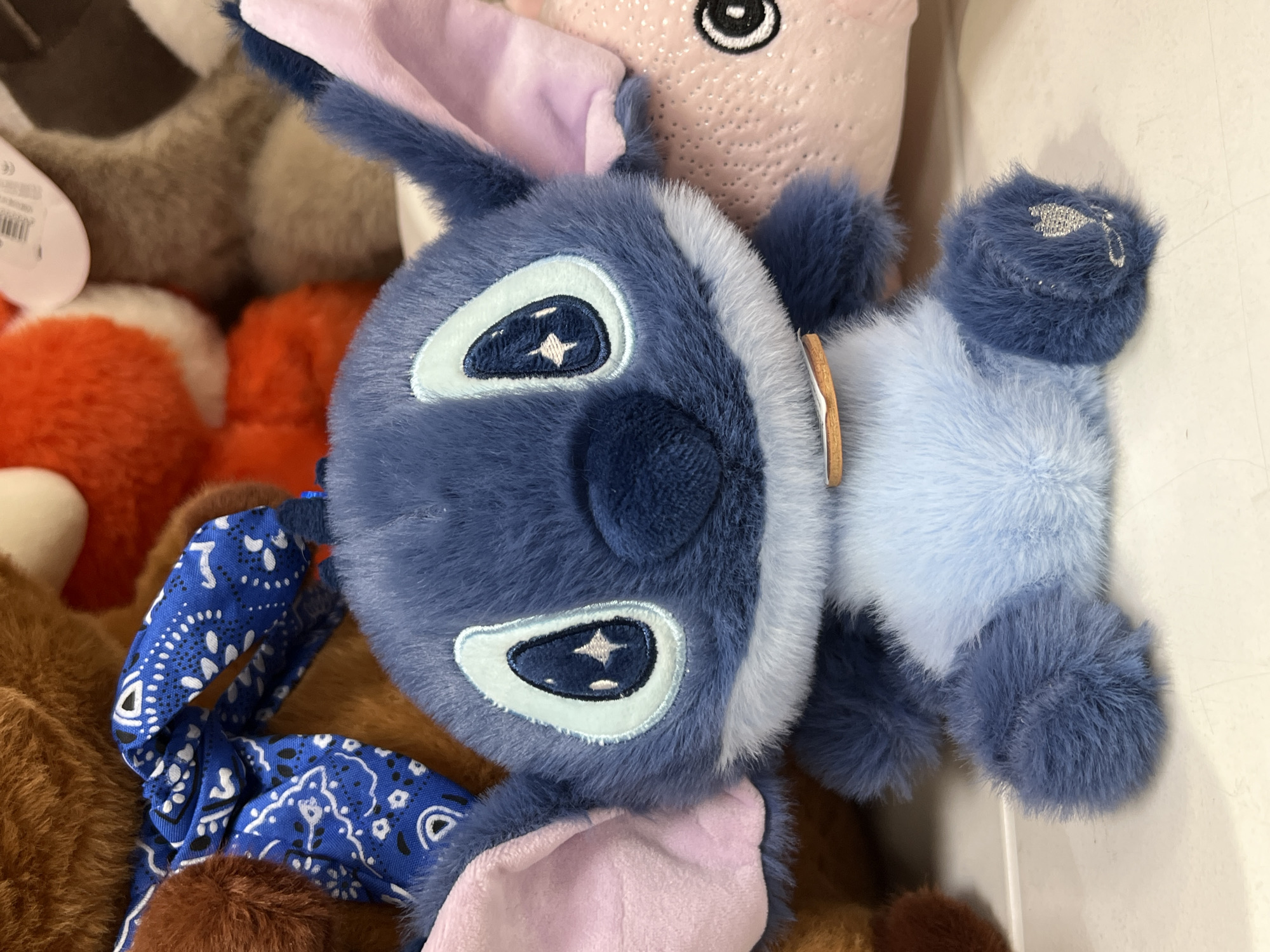 The image shows a close-up of a blue Stitch plush toy sitting on a white surface. The Stitch toy is fuzzy with large eyes that have star-like pupils and pink inner ears. It is wearing a light brown collar. Behind Stitch, there are other plush toys, including a brown bear with a blue bandana tied around its neck and a pink pig-like toy. The background is cluttered with various plush toys of different colors and textures. The lighting is bright and appears to be from overhead. There is no indication of the city of Ridder, Kazakhstan, in the image.