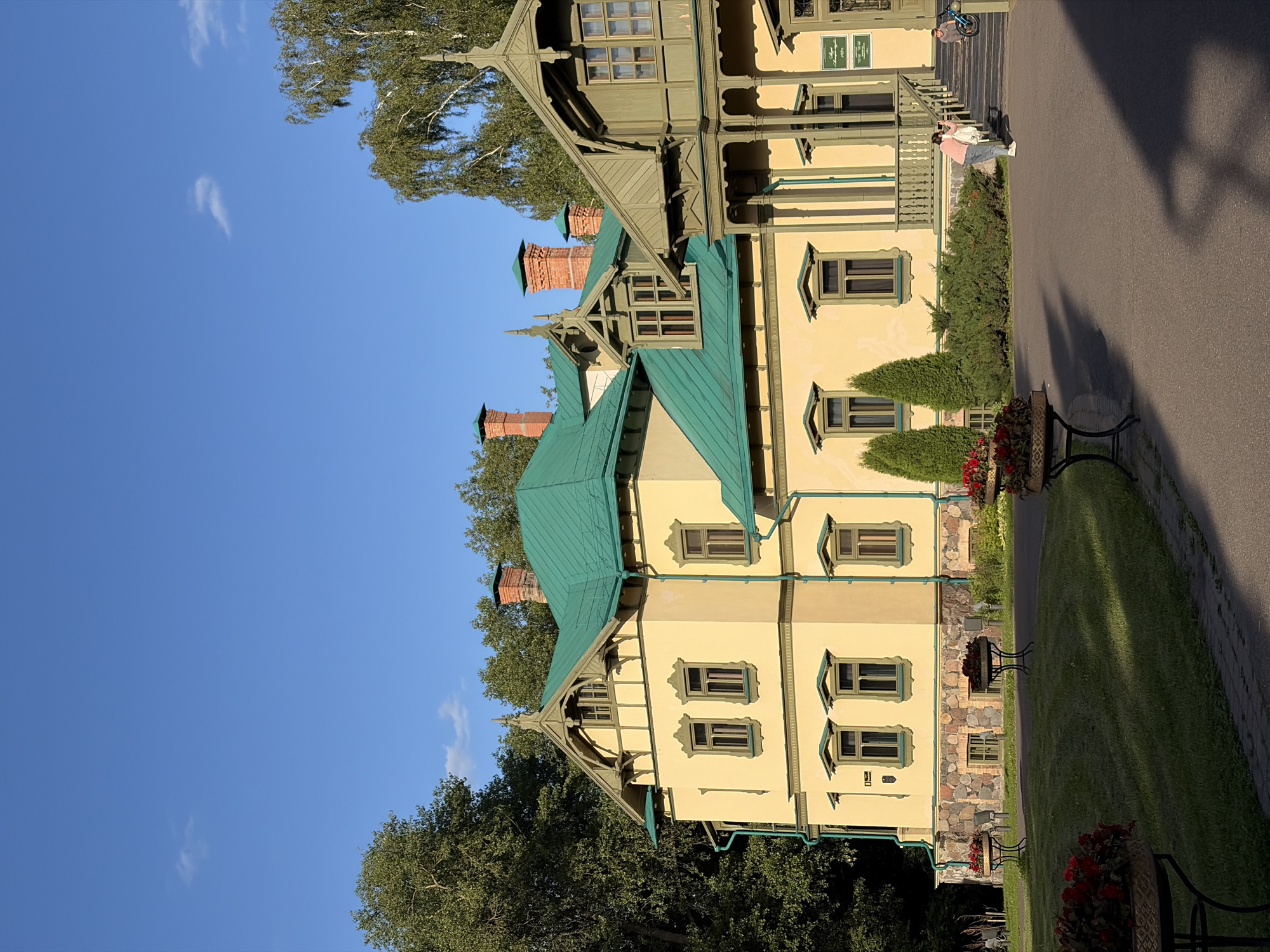 The image captures a charming, historic yellow house with a green roof and intricate wooden trim, set against a clear blue sky. A large, leafy tree is prominent on the left side, with another cluster of trees behind the house. The house itself features multiple windows with decorative green trim, and red brick chimneys rise from the roof. Stone work is visible on the lower portion of the building. In front of the house, a paved walkway runs alongside a lush green lawn with flower arrangements in decorative planters.
Two individuals are visible in the scene. One person, wearing a pink jacket and jeans, is standing on the walkway near the house, appearing to hold a white bag. Another person is further up on a set of stairs, near a bicycle, suggesting movement or arrival/departure. The lighting suggests it is daytime, likely late afternoon due to the long shadows cast on the walkway, indicating warm, sunny weather. The overall atmosphere is peaceful and serene, highlighting the architectural beauty of the house and its well-maintained surroundings. A sign is visible on the upper part of the house, but the text is not legible. The setting is identified as Minsk, Belarus.