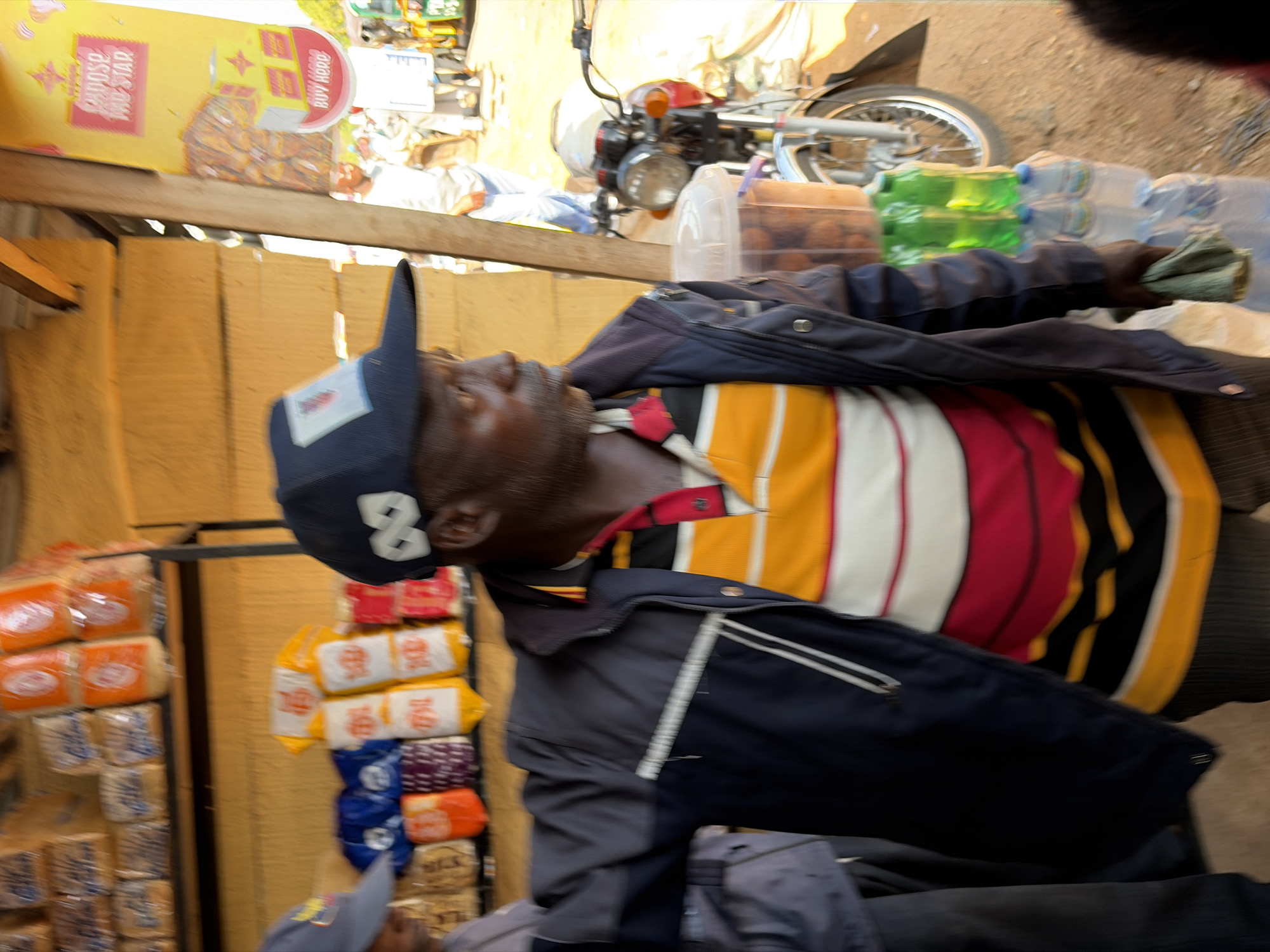 The image shows a person wearing a dark blue baseball cap and a striped shirt with yellow, red, and white bands. They are wearing a dark jacket over the shirt. The person appears to be in an outdoor market stall in Gura Top, Nigeria. Behind the person, shelves are stocked with various goods, including packaged bread or pastries, bottled water, and what appear to be small food items in clear plastic containers. A motorcycle is partially visible in the background, suggesting a bustling street environment. The lighting suggests it is daytime. There is text visible on a yellow sign in the upper left corner which appears to say "CHOOSE THE STAR" and "BUY HERO".