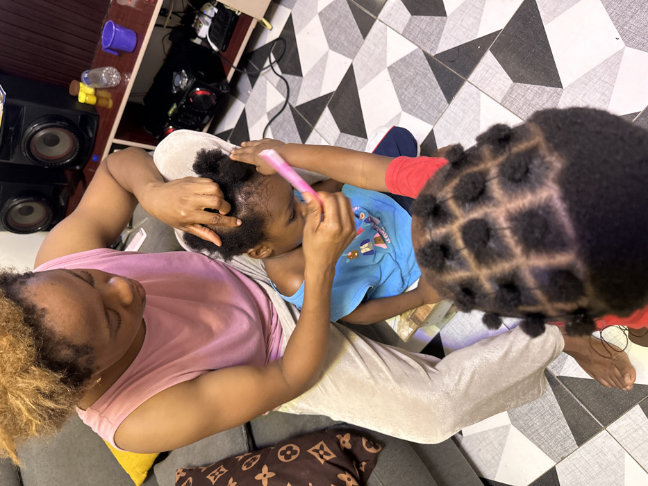 Overhead shot captures a woman in a pink shirt and beige pants, seated and styling a child's hair. The child, in a blue shirt, sits on the woman's lap while someone combs their hair with a pink comb. To the right, another child displays a completed hairstyle featuring a square grid pattern. The setting is a room with a tiled floor, and a glimpse of shelves, speakers, and a pillow with brown and gold designs are visible in the background.