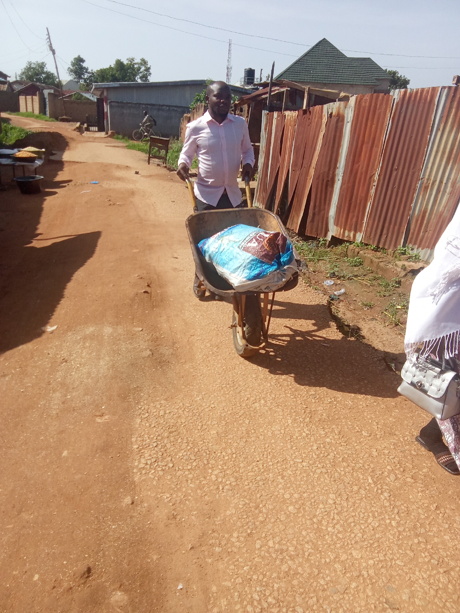 A man in a light pink button-down shirt and dark pants is pushing a wheelbarrow down a dirt road. The wheelbarrow contains a large, blue and white plastic bag. To the man's right, a woman's legs and lower torso are visible. She is wearing a white headscarf, a patterned skirt, and brown sandals. She is carrying a light gray handbag. The road is dusty and uneven, with some gravel. In the background, there are buildings with corrugated iron roofs, a fence made of corrugated metal, and some trees. A power pole and antenna are also visible in the distance. The scene appears to be in a residential area. The location is identified as Jos, Nigeria.