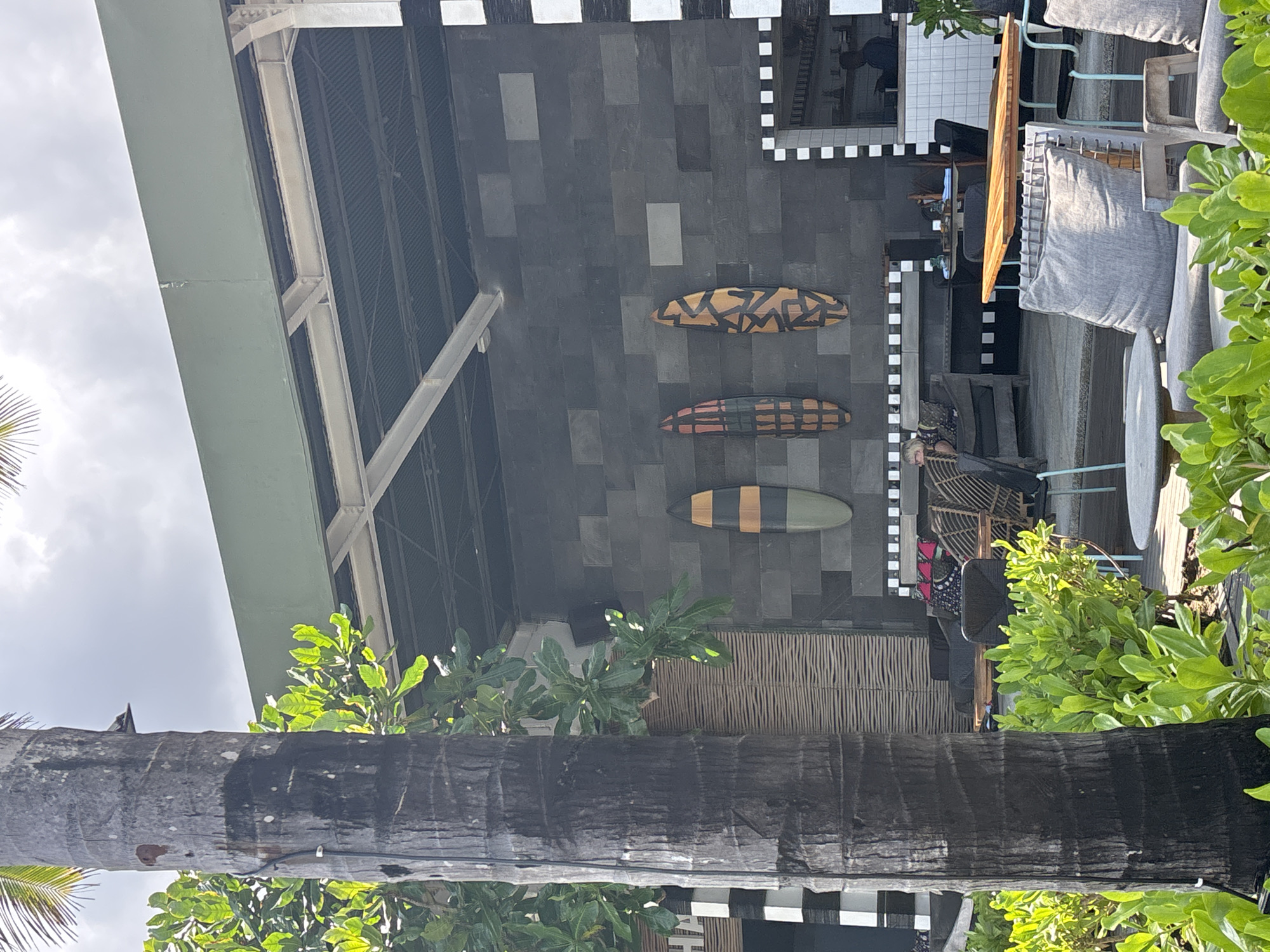 The photograph shows the exterior of a building in North Kuta, Indonesia, as suggested by the local flora. The building has a dark gray, brick-tiled facade, which is decorated with three surfboards of varying designs. The facade features a checkered pattern along the roofline and a section of woven material visible near the bottom of the image. The architecture suggests a modern, semi-open structure, typical of the area. The foreground is dominated by a thick palm tree trunk, which obscures part of the building's lower section. Lush, green vegetation is visible around the building and the palm tree, emphasizing the tropical environment. In the background, a light gray sky is visible. Furnished seating areas are present with wicker chairs and gray cushions. A seated person is partially obscured in one of the chairs.