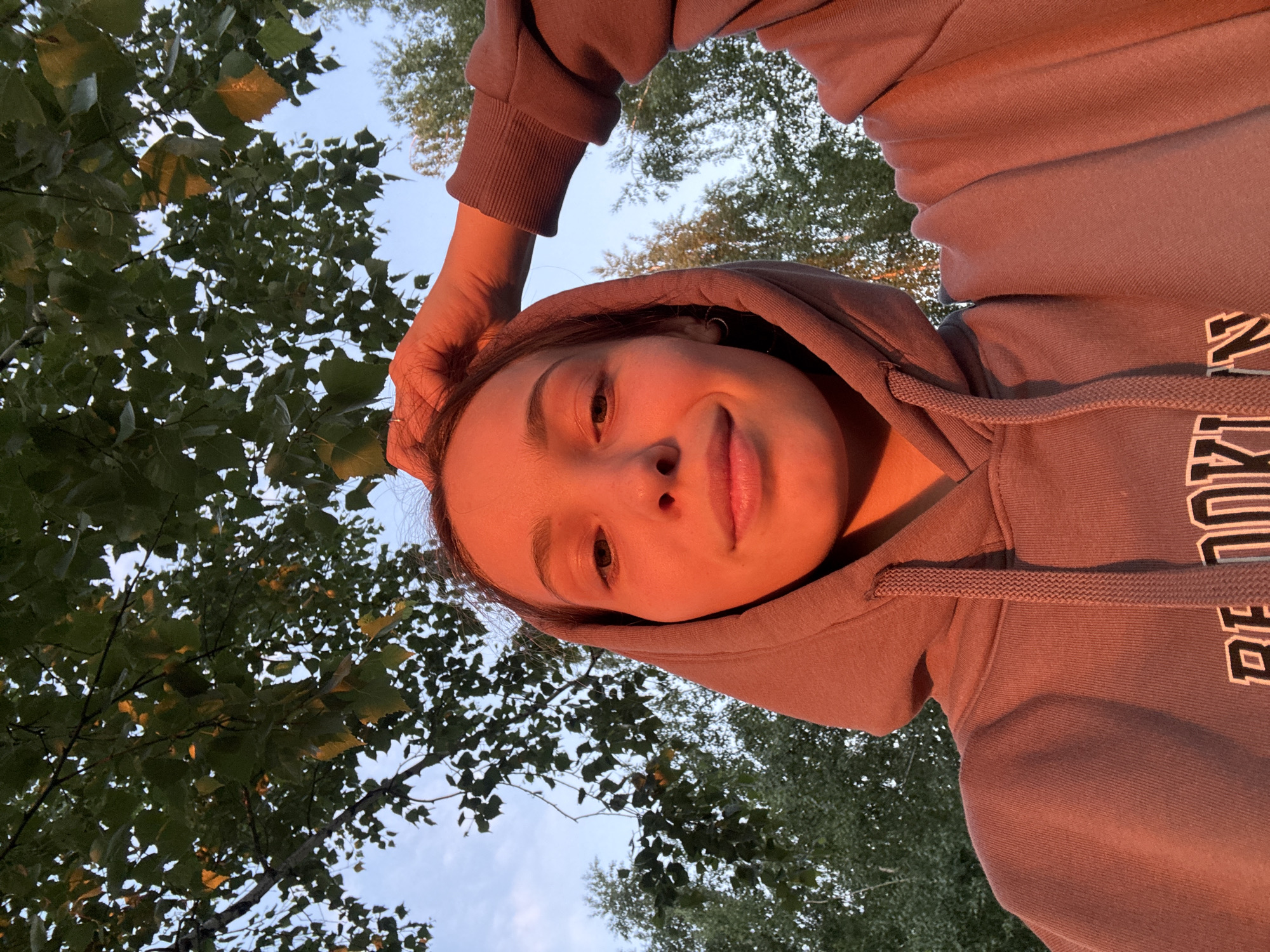 In this waist-up, low-angle selfie, a young Caucasian female with brown hair is positioned centrally against a backdrop of trees and a pale blue sky. She is wearing a muted purple hooded sweatshirt with the hood up. The setting sun casts a warm, orange glow on her face and clothing. Her right hand is raised, touching the top of her head. Visible on the sweatshirt are partially obscured white letters with black outlines, appearing to spell out "BROOKLYN". The environment suggests an outdoor setting, likely a park or natural area within the city of Chaikovsky, Russia, given the foliage and sky.