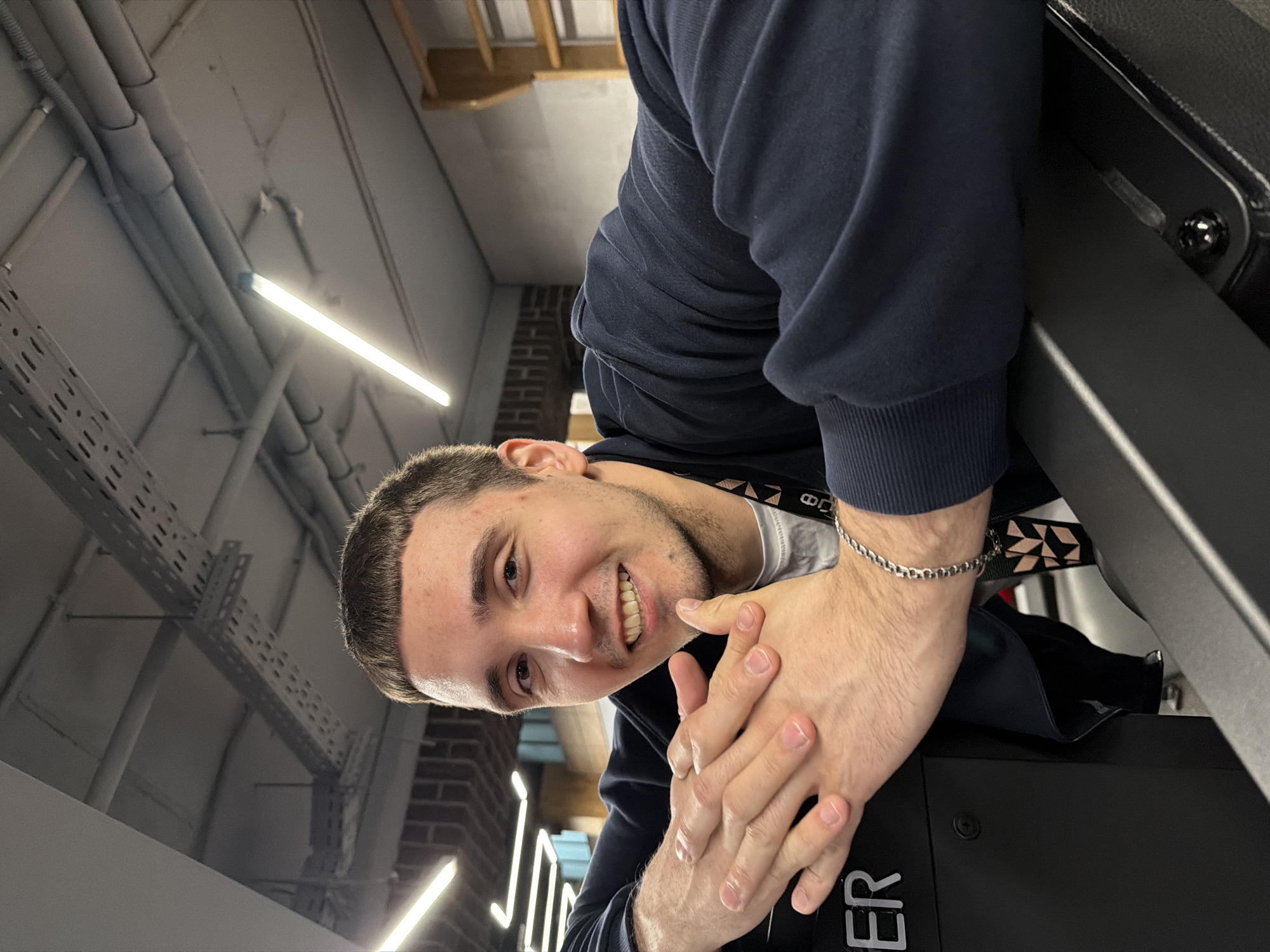A young man is the central subject of this image, smiling warmly directly at the camera. The photograph is taken from a low angle and is significantly rotated counter-clockwise, creating an unusual and dynamic perspective where the man appears to be leaning over or looking down. He has short, dark hair and light stubble, and his expression is friendly and engaging. He is wearing a dark blue long-sleeved top, likely a sweatshirt, over a white t-shirt, and a silver chain bracelet is visible on his left wrist. His hands are clasped together in the foreground, resting on a dark surface.
The setting is an indoor space with an industrial or modern aesthetic. The background reveals a ceiling with exposed infrastructure, including gray pipes and metal cable trays, brightly lit by linear strip lights. A section of a brick wall is also visible behind the man. In the immediate foreground, to the right of the man's hands, is a black piece of equipment or furniture on which the partially visible letters "ER" are inscribed.
The indoor lighting suggests the time of day is not a factor. The man's smile conveys a positive and approachable emotion. No external weather details are discernible. The visible text "ER" is part of a larger word or branding on the equipment. The location, Saint Petersburg, Russia, is provided as context but not visually identifiable within the image itself.