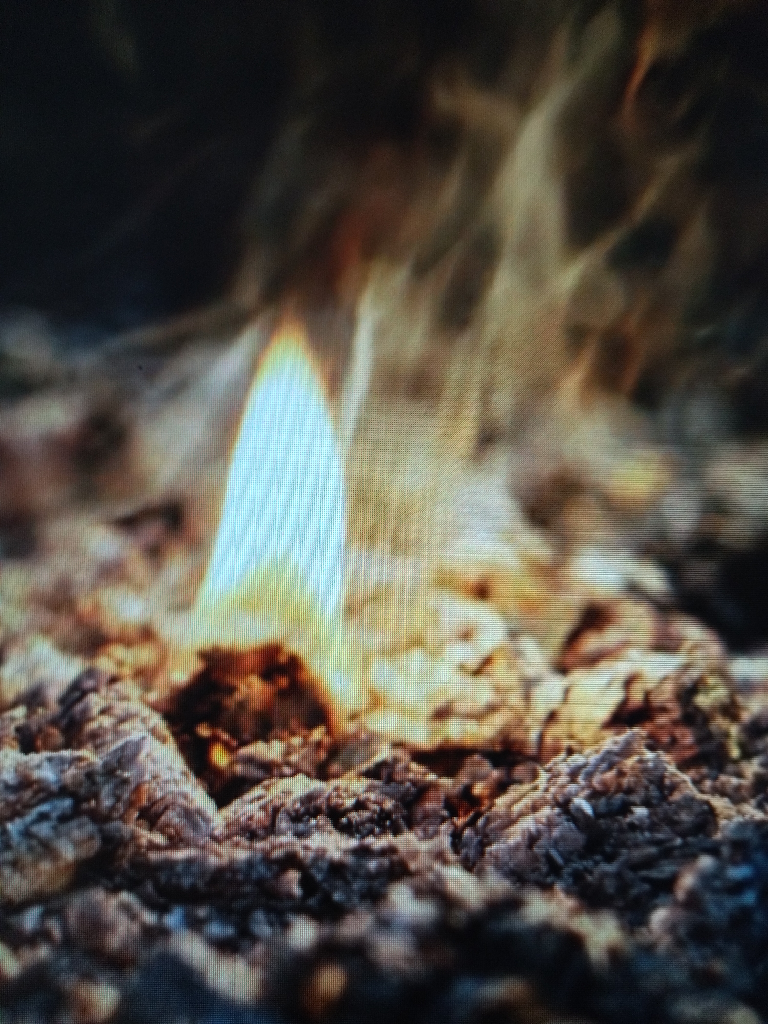 A close-up, shallow depth-of-field, eye-level shot shows a small, bright blue and white flame burning from a pile of ash and embers. The flame is tall and pointed, with a halo of yellow and orange light around it. The ash and embers are dark gray and brown, with some reddish highlights. The background is dark and out of focus, with wisps of orange and yellow smoke or flame visible. The image appears to have been taken with a low-resolution digital camera or is a photograph of a screen.