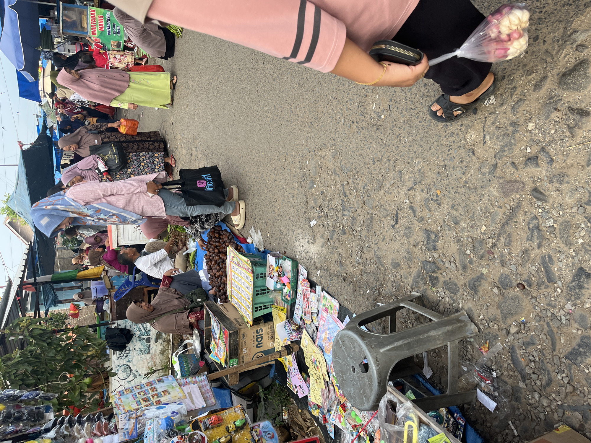 The image depicts a bustling outdoor market scene in Kemuning, Indonesia, likely a Sunday market given the crowd and variety of goods. The market is set up along a paved road, with stalls lining one side and people milling about.
In the foreground, a woman in a pink shirt and black pants is visible from the waist down, holding a small black purse and a plastic bag containing a bundle of shallots. She is wearing black sandals. To her left, a grey plastic stool sits on the rough, cobblestone-like pavement, surrounded by scattered debris and some children's toys.
Further into the market, a woman wearing a brown hijab and a long brown dress is seated behind a display of goods, which includes what appear to be notebooks and stationery. Next to her, a pile of brown nuts or seeds is visible, alongside a green plastic basket filled with similar items. To the left of the seated woman, there are more colorful children's books and toys spread out on a black tarp.
Other individuals in the scene are seen browsing and interacting with the stalls. Many women are wearing hijabs in various colors, including light blue, pink, and beige. One woman in a light blue hijab and jeans is carrying a black tote bag with "Mcory" written on it. Another woman in a lime green dress and a pink hijab stands near a stall that appears to be selling food, indicated by a sign that reads "MARTABAK MANIS". The stalls are covered with various tarps and awnings to provide shade. The general environment suggests a vibrant, everyday market activity.