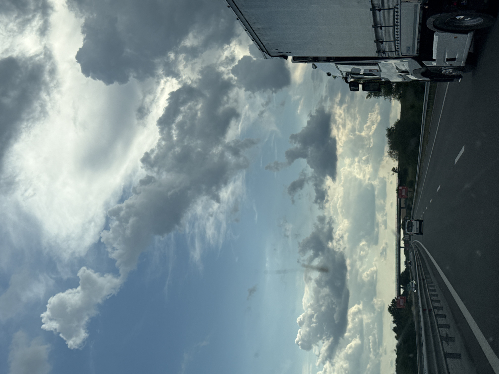 The image depicts a view from a vehicle traveling on a highway. The sky is filled with a dramatic mix of dark, heavy clouds and patches of bright blue sky. In the foreground, a white truck is partially visible on the right side of the frame, traveling in the same direction. The highway stretches ahead, with other vehicles, including a white van and a car, visible in the distance. The road is bordered by safety barriers and greenery on the left. An overpass is visible in the far distance. The overall impression is one of a journey under a dynamic and changing sky. The location is specified as the city of Yazdach, Bulgaria.