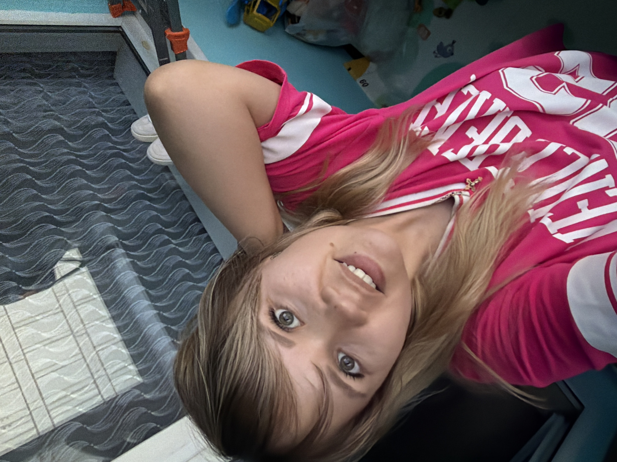A young woman is posing for a selfie. She is wearing a pink t-shirt with white lettering. She has fair skin, light brown hair, and gray eyes. She is smiling slightly. The background includes a patterned floor, a blue wall, and toys. The location is Chaikovsky, Russia.