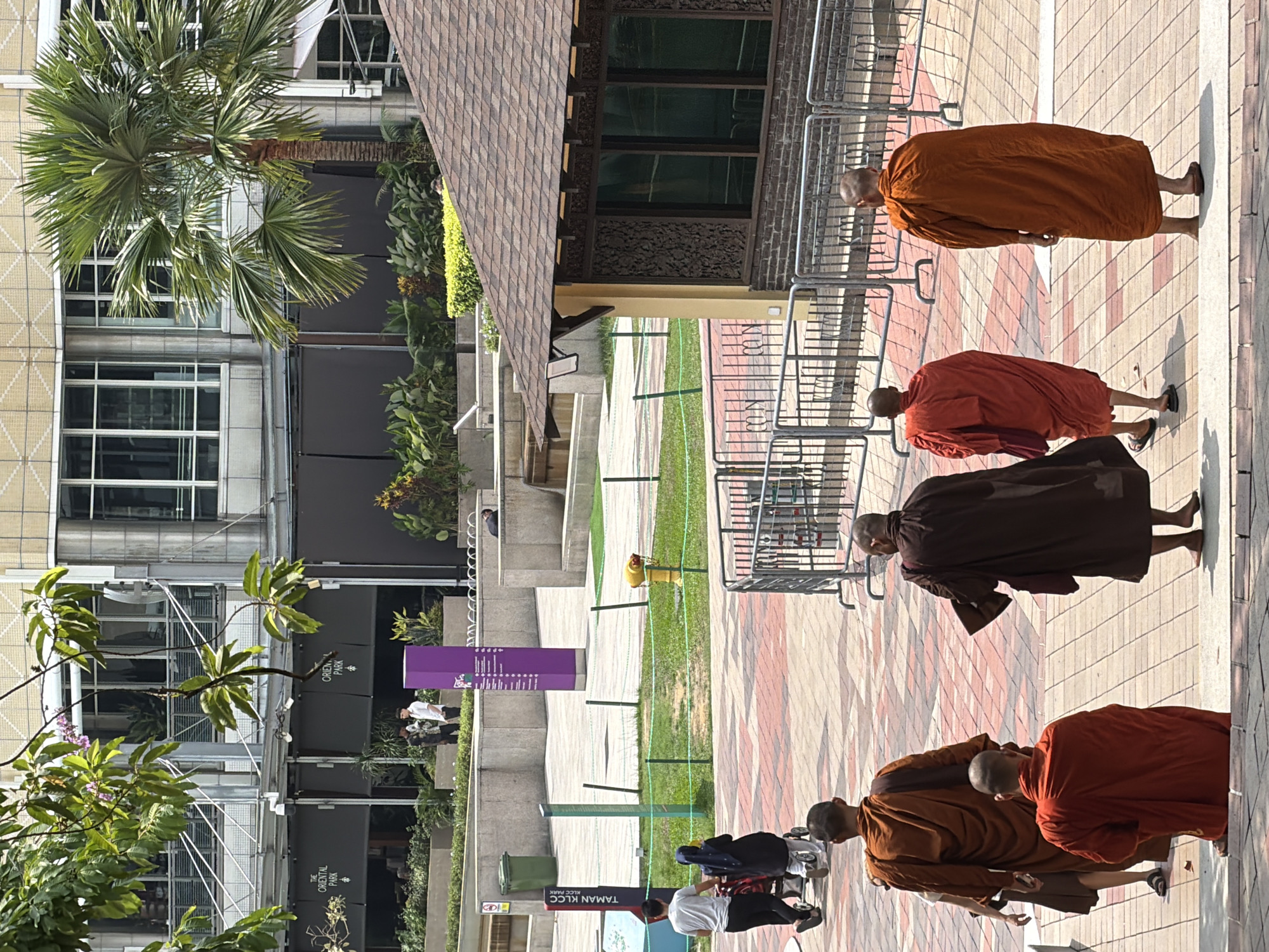 The image depicts a scene in Kuala Lumpur, Malaysia, potentially in or near the KLCC Park. The environment includes modern buildings, trees (including palm trees), a tiled walkway, and a green grassy area separated by metal railings. Several Buddhist monks wearing orange and brown robes are walking along the tiled walkway. Other individuals, including people dressed in casual attire, are also present, suggesting the location is a public space. Signage in Bahasa Malaysia is visible, supporting the location within Malaysia.