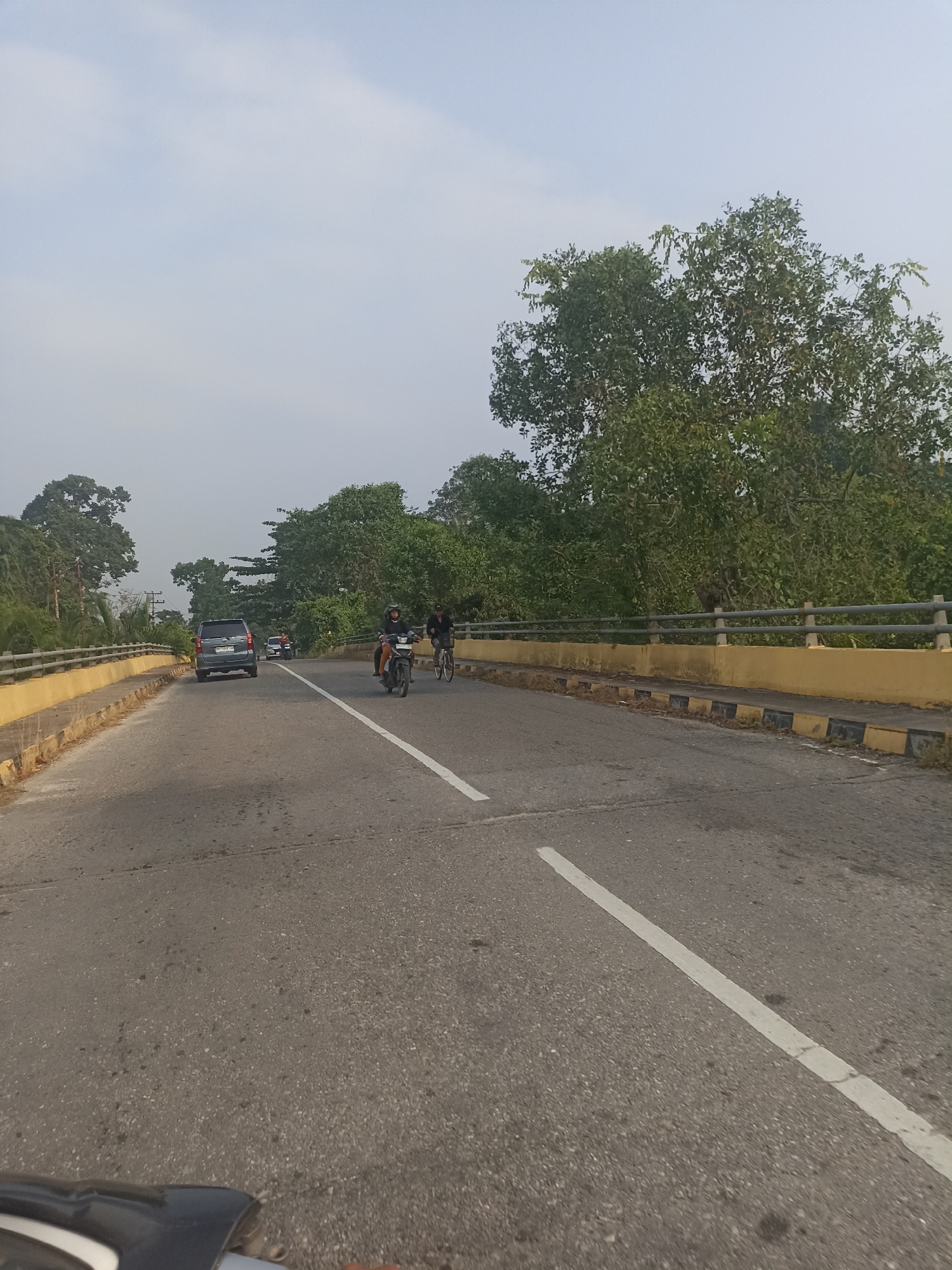 The image shows a two-lane asphalt road, likely a bridge, with a yellow barrier and metal railing on the left and right sides. Lush green trees line the sides of the road. The sky is overcast with a pale blue hue.
In the foreground, on the right lane, a person is riding a motorcycle, wearing a helmet. To their right, a person is riding a bicycle.
Further down the road, on the left lane, a dark-colored SUV is driving away from the camera. Behind the SUV, there are other vehicles visible, including a motorcycle.
The location is identified as Kampung Tengah, Indonesia. The scene depicts moderate traffic on a road surrounded by vegetation.
