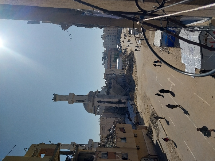 This image depicts a wide-angle view of a heavily damaged urban street scene in Khan Yunis, Palestinian Territory, centered on the Al-Farouq Mosque. The mosque, situated in the mid-ground, shows significant structural damage to its lower sections and surrounding buildings, though its tall, cylindrical minaret and central dome remain largely intact.
To the left, a multi-story building exhibits extensive destruction, with large sections of its facade collapsed, exposing internal rooms and reinforced concrete elements. Piles of rubble and debris are scattered across the ground adjacent to this structure.
The foreground features a paved road, partially covered with sand and dirt, with white directional arrows painted on its surface. Approximately 15 to 20 individuals, appearing as civilians in dark and light clothing, are observed walking along the street and in the distance, casting long shadows to the left, indicative of a bright morning or late afternoon sun.
In the lower right foreground, the edge of a building or wall made of reddish-brown material is visible, partially obscured by a section of a wire fence, likely barbed wire. Below this, a makeshift structure, constructed with white and blue tarpaulins, stands on sandy ground next to the road. The sky is clear and blue, with a prominent sun flare originating from the upper right quadrant.