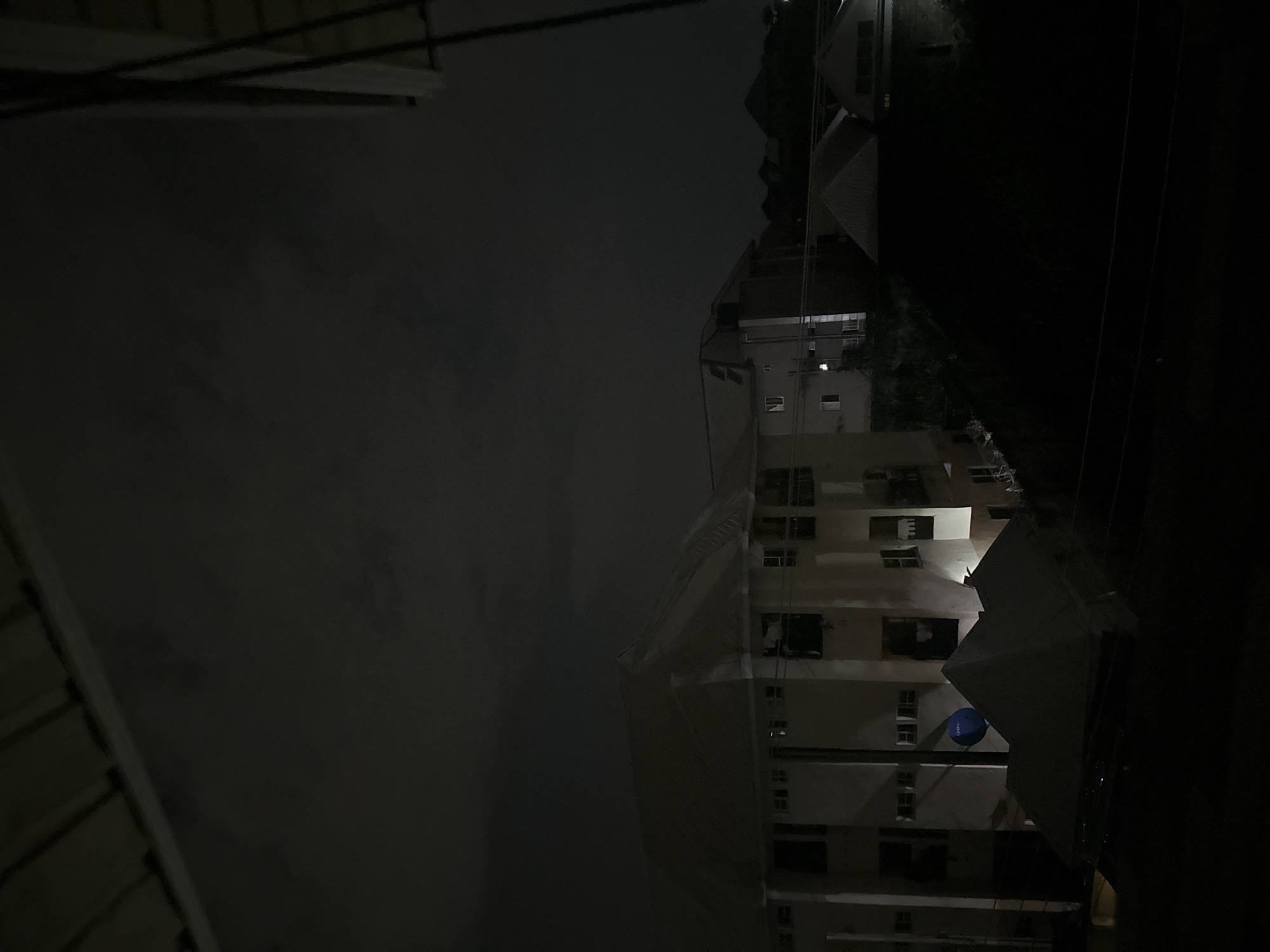 The image depicts a nighttime scene in Ihiagwa, Nigeria, featuring a partially illuminated, multi-story building under construction or renovation. The building's facade is visible, showing multiple window openings and balcony structures. Scaffolding covered in white tarpaulin or protective sheeting is present on one side of the building, partially obscuring the view. The sky is dark and cloudy. The area immediately surrounding the building is dark, with some hints of vegetation on the right side of the frame. Overhead wires are visible against the dark sky.