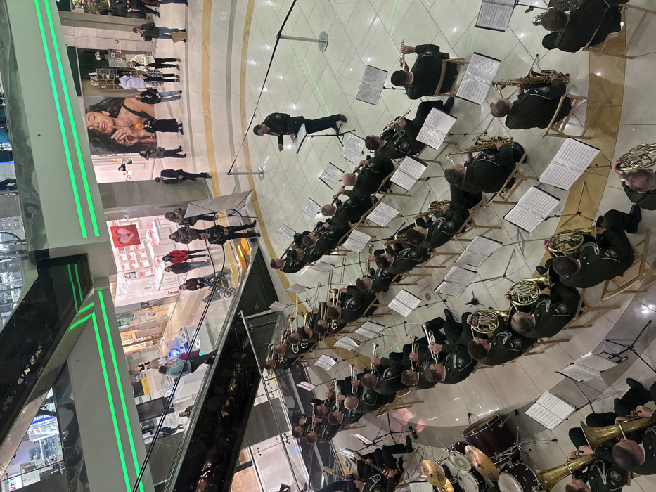 A military-style band, consisting of approximately 30-35 male musicians in olive-green uniforms, performs an indoor concert. The musicians are seated on wooden chairs, each with a music stand, playing a variety of brass instruments (trumpets, trombones, French horns), woodwinds (saxophones, clarinets, flutes), and a drum kit. A male conductor, in a matching olive-green uniform, stands before the ensemble at a central music stand, actively directing.
The event takes place in the central atrium of a multi-story modern shopping mall, specifically identified as the Blockbuster Mall in Kyiv, Ukraine. The performance area is on the ground floor, characterized by light-colored polished flooring and partially cordoned off by a dark rope barrier. Approximately 15-20 spectators, including adults and children in civilian attire, are observing the performance from behind the barrier and from upper levels. One individual is visible descending an escalator.
The mall's architecture features multiple levels with glass railings and several escalators. Distinctive horizontal green neon light strips illuminate the escalator structures and overhead architectural elements. Retail storefronts with illuminated product displays and large promotional posters, one depicting a female model, line the perimeter on multiple floors surrounding the atrium.