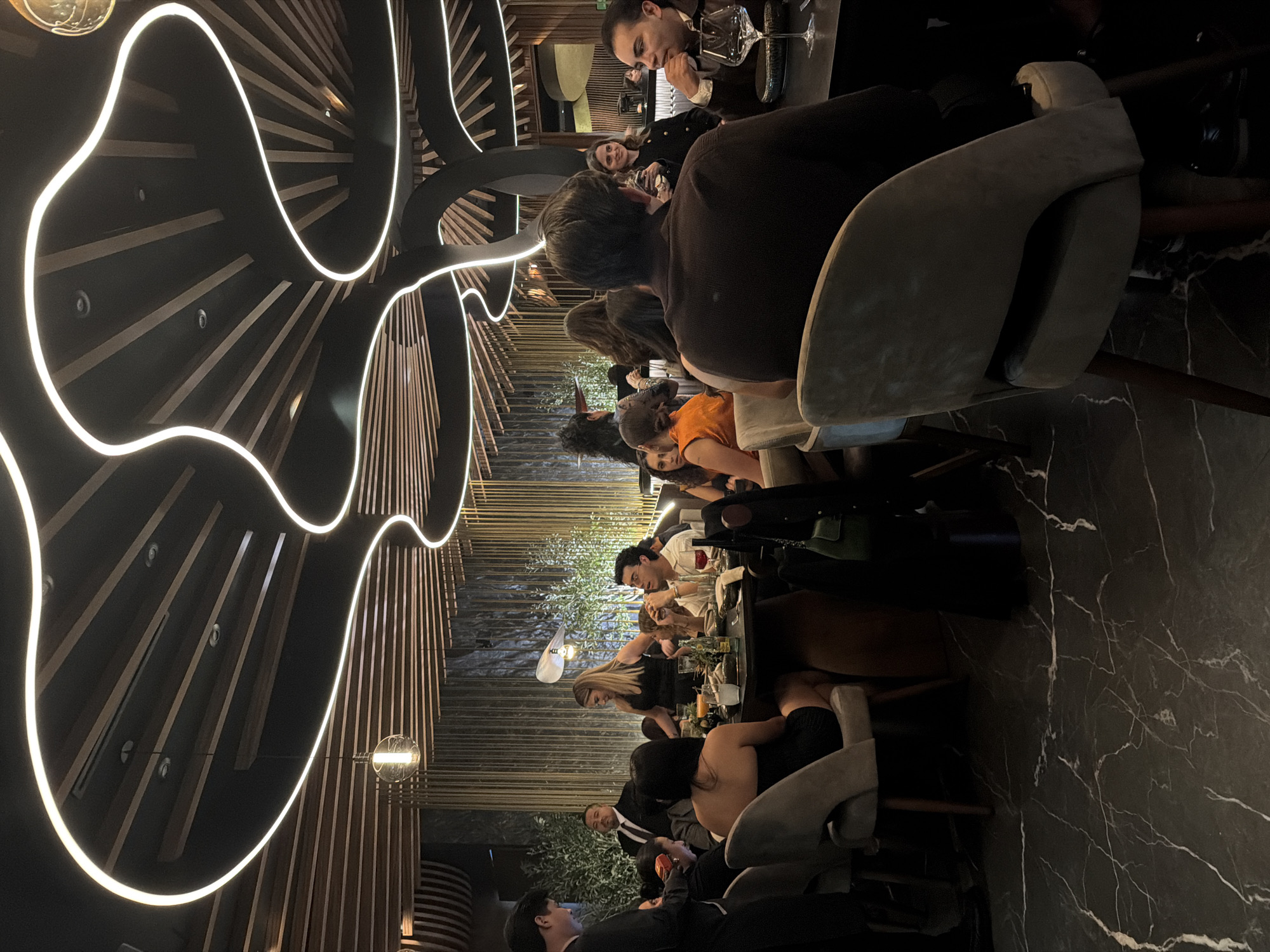 The image shows a restaurant interior. The scene appears to be a gathering of people seated at tables, possibly dining or socializing. There are numerous individuals, including men and women of various ages. Several are engaged in conversations, while others seem to be eating or drinking. The restaurant features a distinctive ceiling design with dark panels interspersed with light-colored elements that create a pattern. The walls are adorned with wooden slat-like decorations. The location might be in San Bernardino Tlaxcalancingo, Mexico, though this is inferred without concrete visual proof.