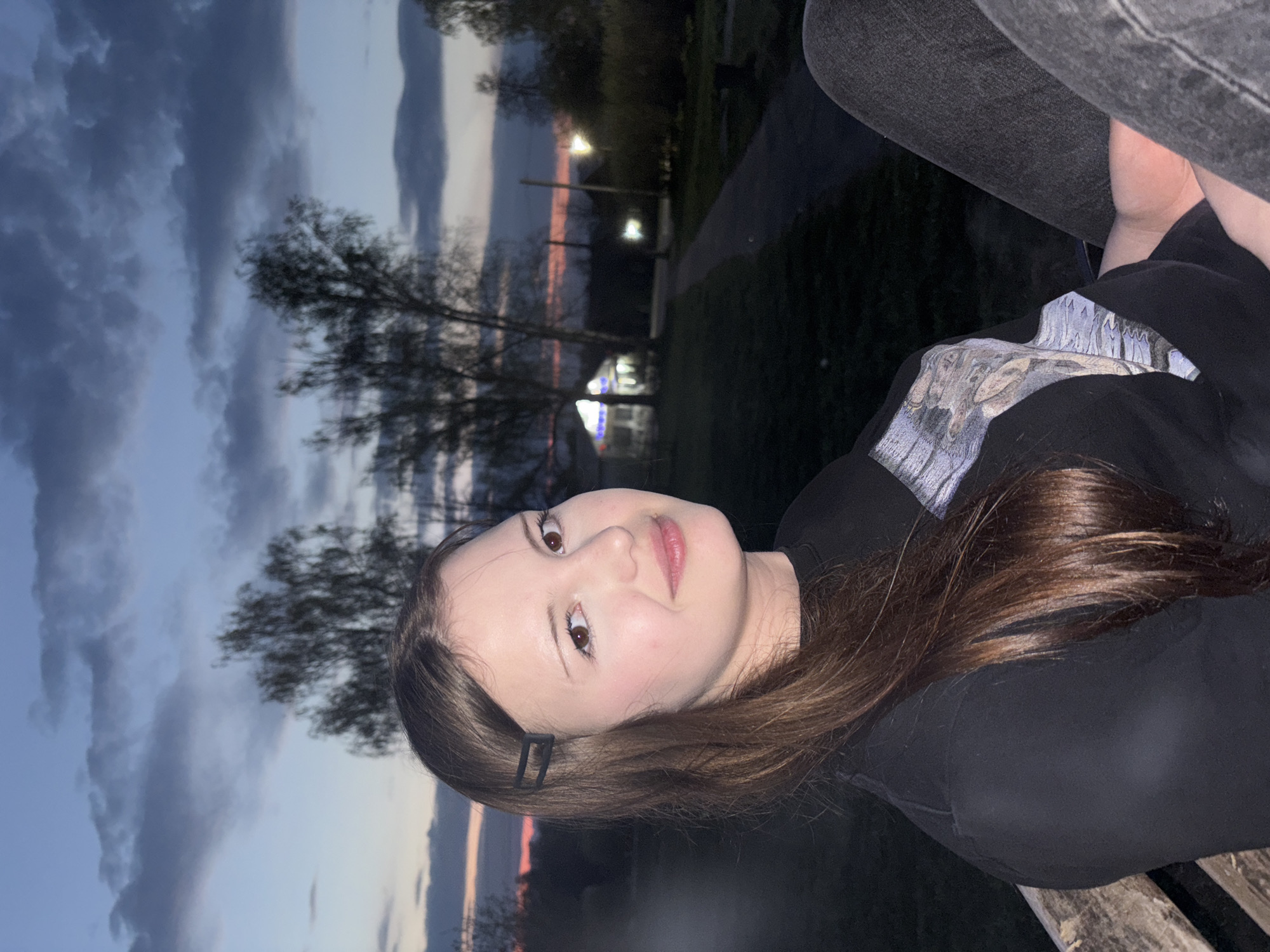 A young woman is pictured outdoors in what appears to be a park or green space near Фоки, Russia. She is wearing a black top and jeans and has medium-length brown hair with a black clip. In the background, there are trees and a sky with clouds illuminated by the setting sun. In the distance, there are lights, possibly from buildings or street lamps.