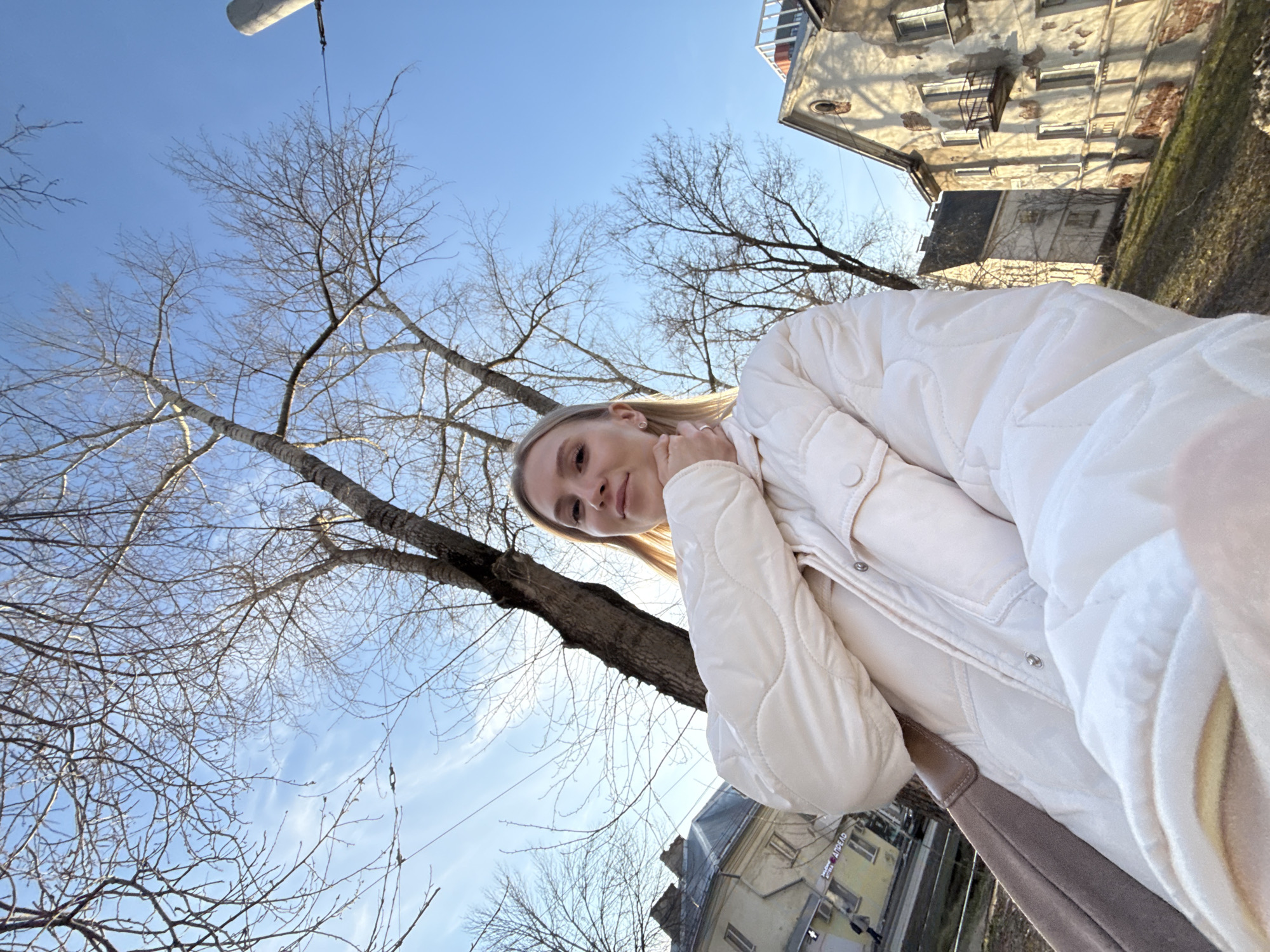 A woman in a white jacket takes a selfie outdoors on a sunny day. Bare trees fill the sky in the background. A building is partially visible in the background. The location is Izhevsk, Russia.
