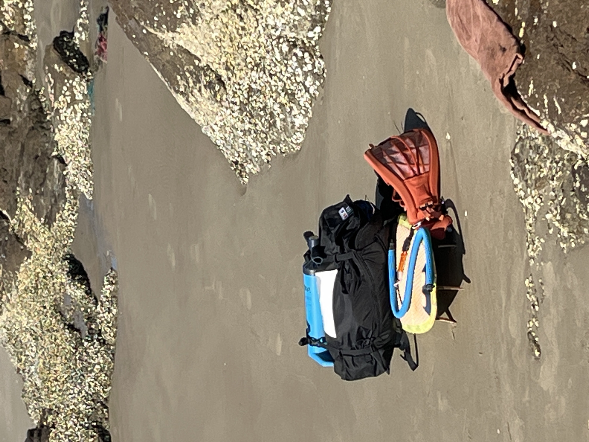 The image shows a beach scene in Mũi Né, Vietnam. In the foreground, there are several objects on the sand. A large black backpack with a blue and white top section is prominent. Next to it, a small yellow and blue object, possibly a child's surfboard or bodyboard, is propped up by wooden sticks, with a blue tube coiled on top. To the right of these items, an orange bag or container with a mesh-like design is lying on its side. A piece of brown cloth is visible on a rock in the lower right corner. The background features sand, water, and rocks covered in small white shells. Footprints are faintly visible in the wet sand.