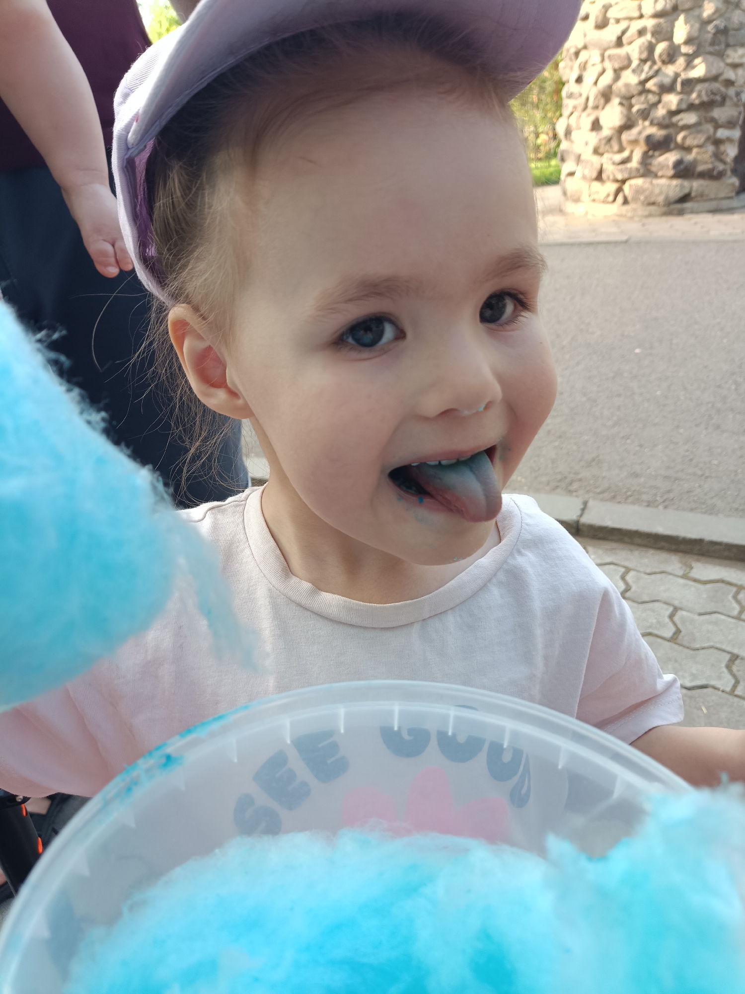 A young child with light skin, wearing a light-colored t-shirt and a light purple cap, is eating blue cotton candy. The child's tongue is blue from the candy. The cotton candy is in a white bucket with pink and blue decorations. The child is being held by an adult, visible only by their arm and shirt. In the background, there is a stone structure and paved ground. The location is in the city of Chaikovsky, Russia.
