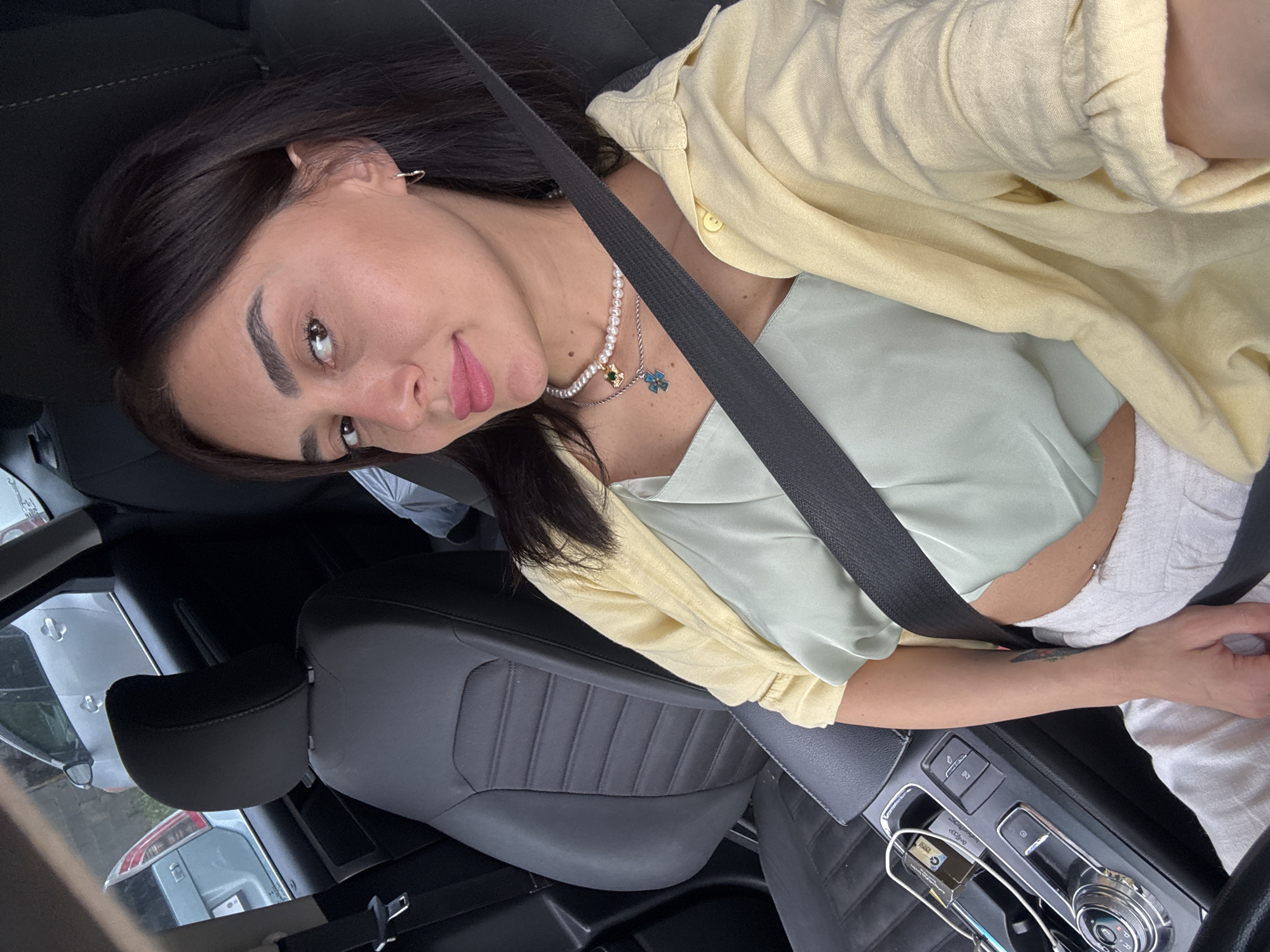 A woman with dark hair, wearing a light yellow open-collared shirt over a light green camisole and white pants, is seated in the driver's seat of a car. She has a seatbelt fastened across her chest. She is looking towards the camera, with makeup on, including pink lipstick. She is wearing two necklaces, one with pearls and a pendant, and small hoop earrings. A tattoo is visible on her left forearm. The car's interior is visible, including the black leather seats, a center console with a gear shifter, and part of the steering wheel. Outside the car, other vehicles are visible through the windows. The scene is consistent with being in the city of Tbilisi, Georgia, given the typical car models and urban environment often found there.