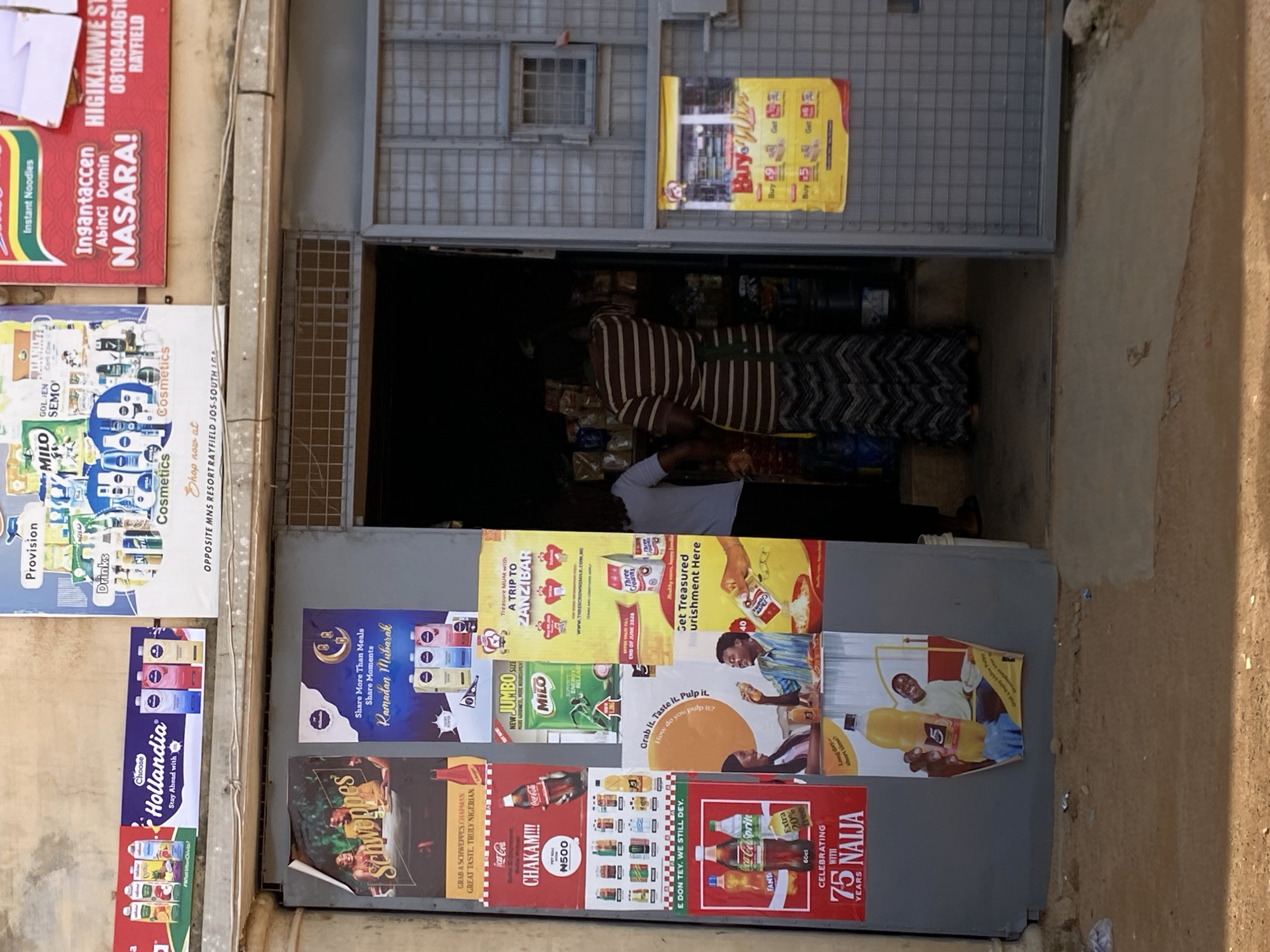 The image depicts the exterior of a small shop in Jos, Nigeria. The shop entrance is open, revealing shelves stacked with various goods inside. A woman in a striped dress and apron stands behind the counter within the shop. Outside the shop, the walls are adorned with numerous colorful advertisements for beverages, food items, and cosmetics. Prominent among these are posters for Hollandia, Schweppes, Coca-Cola, and Milo. Some of the advertisements are in local languages. The ground in front of the shop is a dusty, uneven surface. The building appears to be made of concrete. The overall scene suggests a typical Nigerian street-side convenience store.