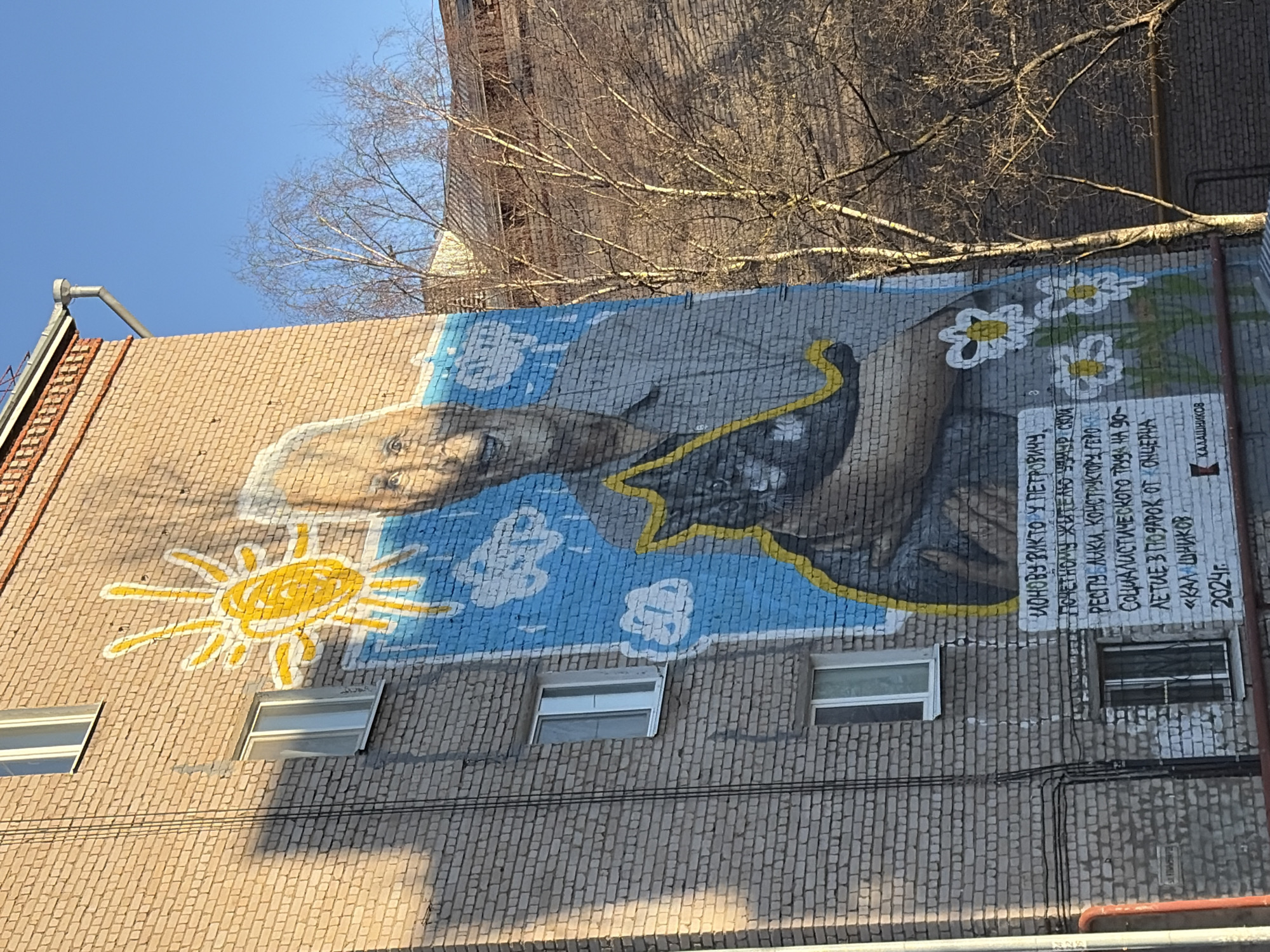 A mural of a man in a blue sky is painted on a brick building. Below the mural are windows. A yellow sun is painted to the left of the windows and flowers are painted to the right of the mural. The mural appears to be dedicated to a person named "Monov Victor U Petrovich" and the Kalashnikov company. The building is located in Izhevsk, Russia.
