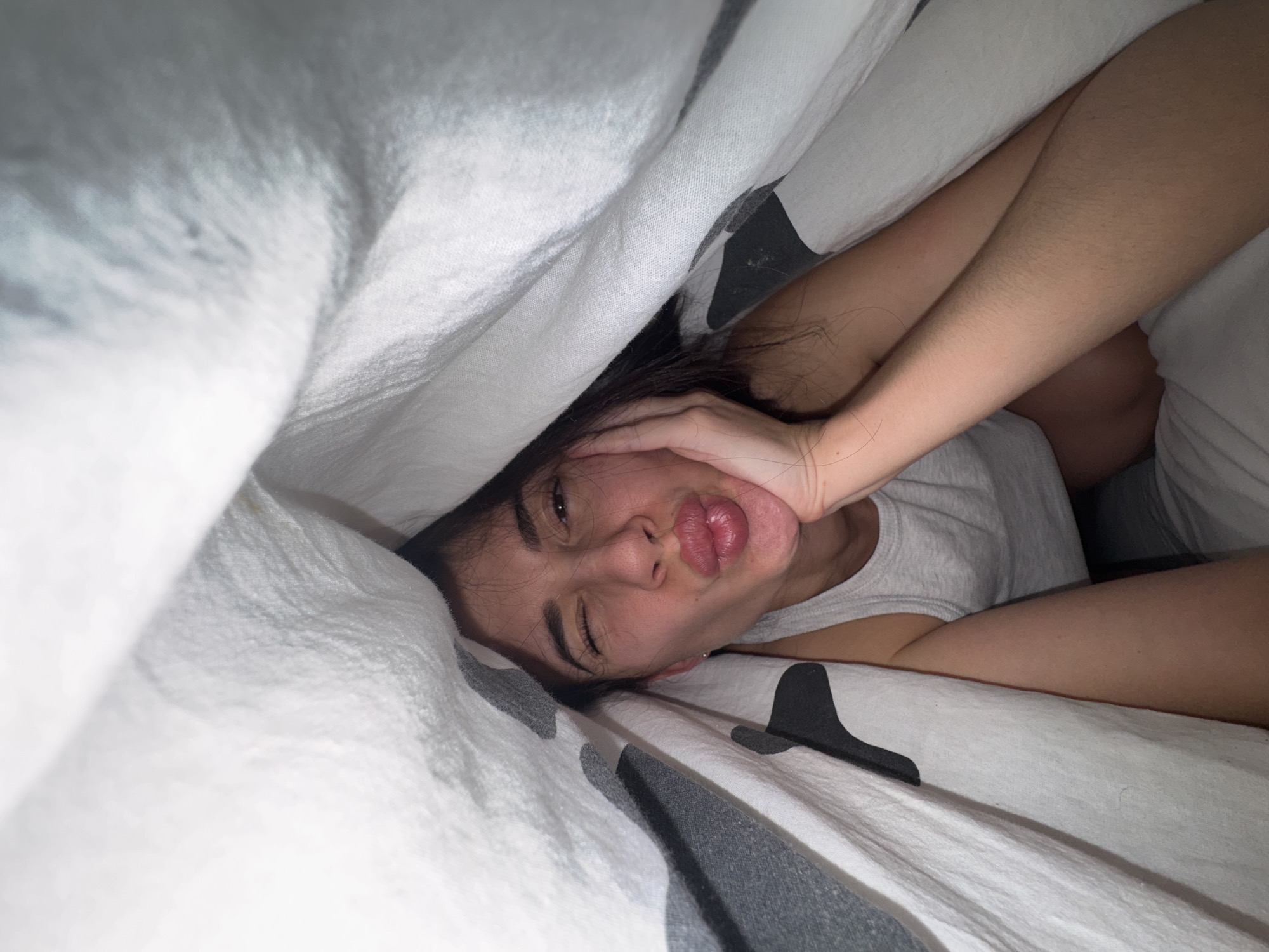 A person lies in bed under white and gray patterned sheets and a white duvet. The person, likely a young woman, is making a pouting face, with one eye closed and their left hand resting on their cheek. Part of her dark hair is visible. She is wearing a gray shirt. A black object, possibly a hair clip, lies on the bed. The location is unknown.