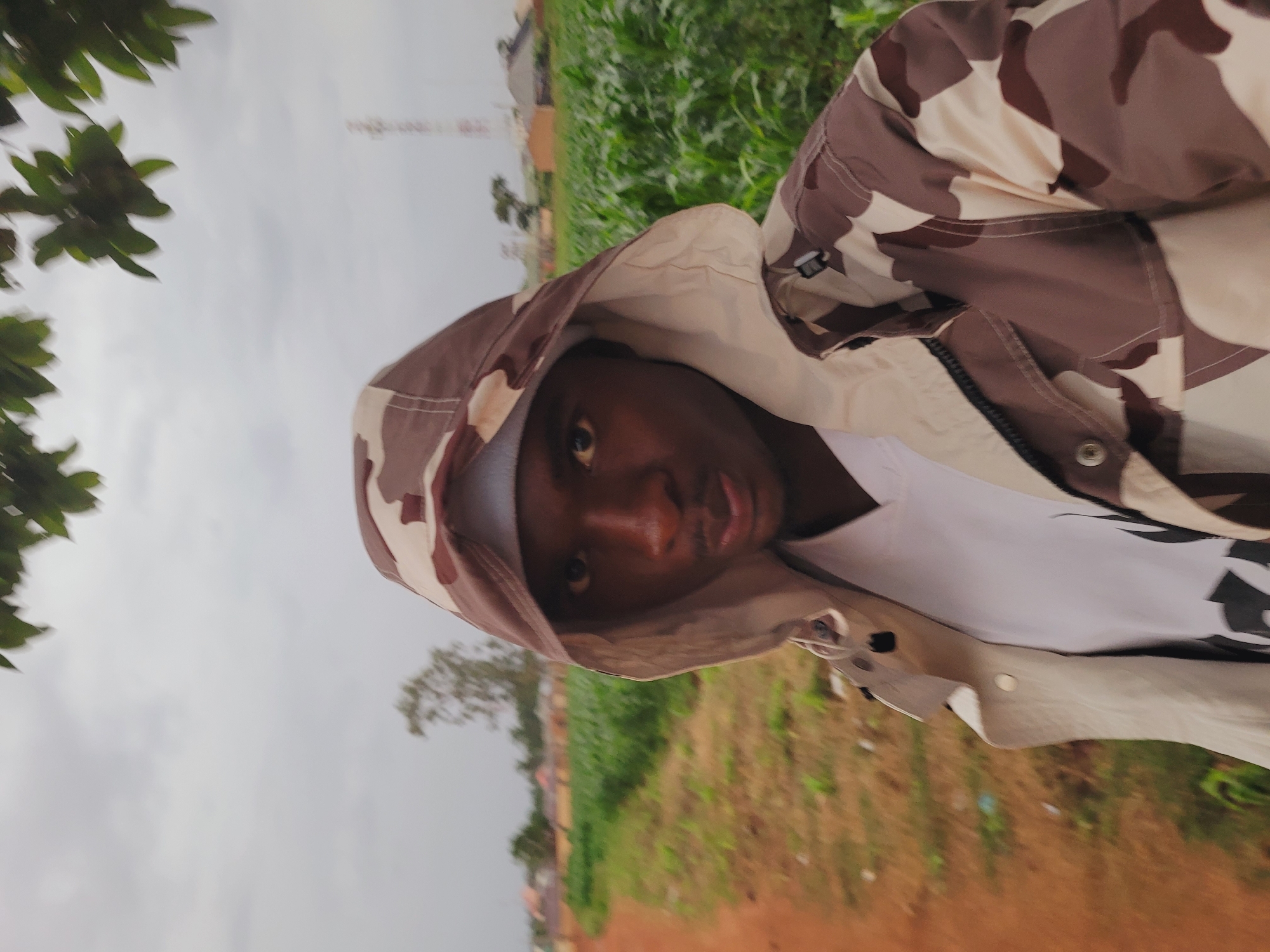 The image, correctly oriented, captures a person with dark skin looking directly at the camera. They are wearing a hooded jacket featuring a brown, beige, and off-white camouflage pattern, pulled up to cover their head. A white t-shirt with a dark graphic design on the right side is visible beneath the jacket. The person's expression is neutral, and their dark eyes engage the viewer.
The scene is outdoors, set under an overcast, light grey sky. In the upper left corner, vibrant green foliage from a tree or bush is visible, slightly out of focus. The background to the right and bottom of the frame shows expanses of green grass or crops and reddish-brown earth, suggesting a natural or agricultural environment. In the mid-distance, behind the person's left shoulder, some indistinct, possibly reddish-brown buildings or structures can be discerned.
The person appears to be posing for a photograph. The cloudy sky indicates an overcast day, resulting in soft, diffused lighting. The overall setting, combined with the provided location context, points to an outdoor scene in Bukuru, Nigeria. No discernible text is visible on the clothing or in the background.