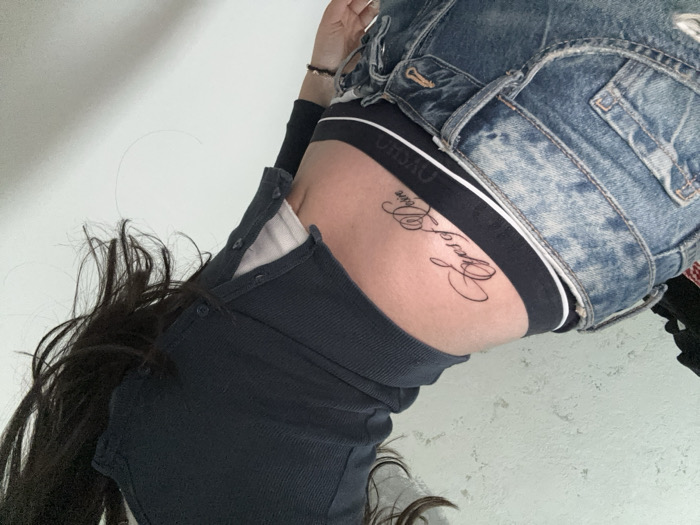 The image depicts a close-up, angled view of an individual, contextually understood to be within an indoor setting in Yerevan, Armenia. The subject, identifiable by long, dark hair at the top, is partially clothed. They wear faded blue denim jeans, unfastened at the waist, revealing the upper portion of dark-colored underwear with a white stripe and text, partially visible as 'OVSHO'. A dark blue, long-sleeved, ribbed top with button detailing is worn over a white undershirt or bra. A prominent black script tattoo, reading "Eyes of Desire", is visible on the lower right back/hip area, positioned above the underwear line. The background consists of a plain white wall on the right and a textured, lighter grey wall on the left. Dark, indistinct fabric is visible in the upper left corner.