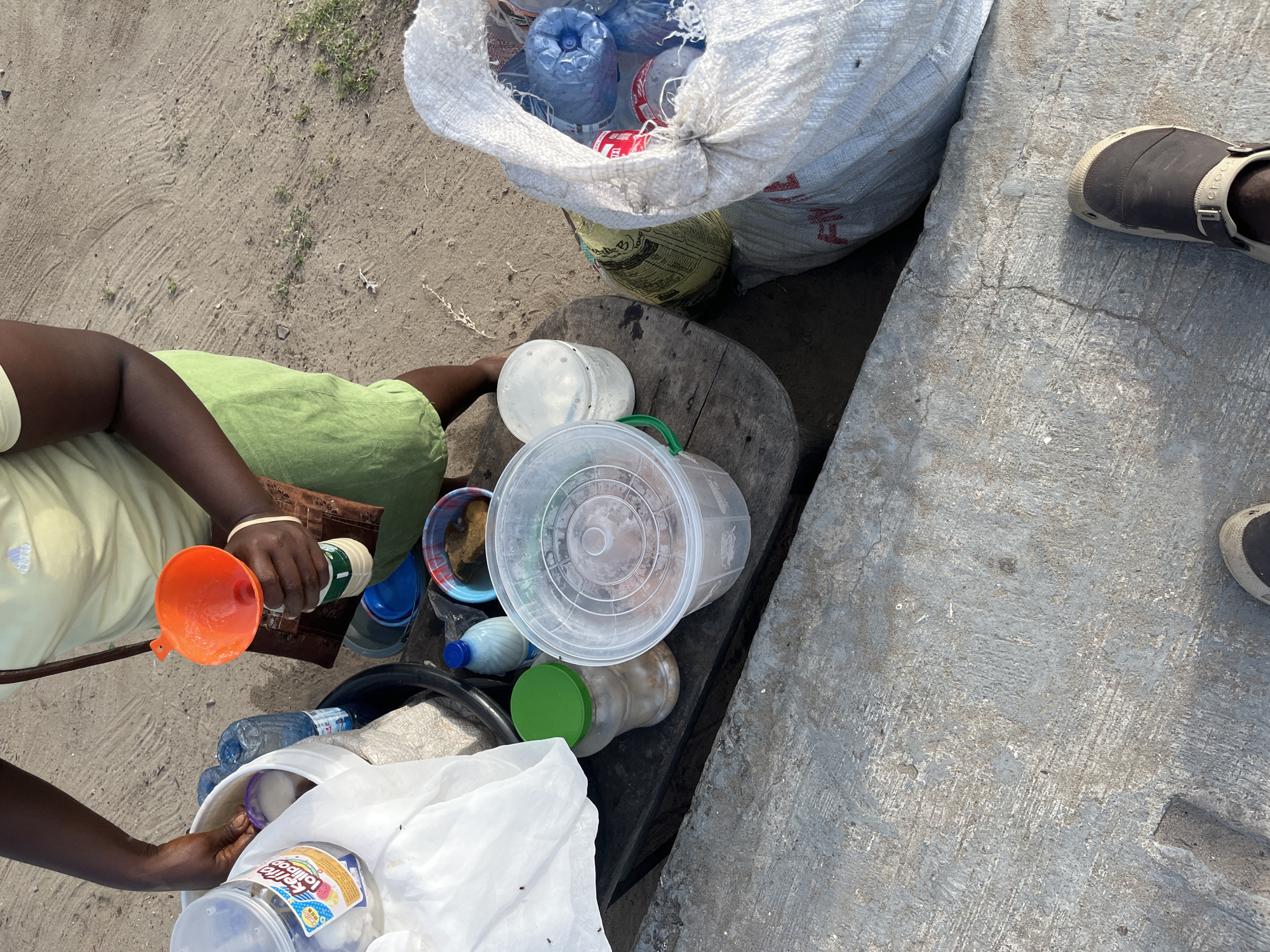 From an elevated, perpendicular perspective, a person is shown sorting items on a makeshift table or stand in Badagry, Nigeria. The individual wears a lime green top, khaki pants, and a brown wrist band. They are holding an orange funnel in one hand and pouring the contents of a bottle into it. The materials being sorted include various plastic bottles, containers, and bags. A large white bag, filled with plastic bottles, is positioned next to the work area. The surrounding environment appears to be a concrete surface. A pair of brown clogs is visible. The setting is outdoor under daylight.