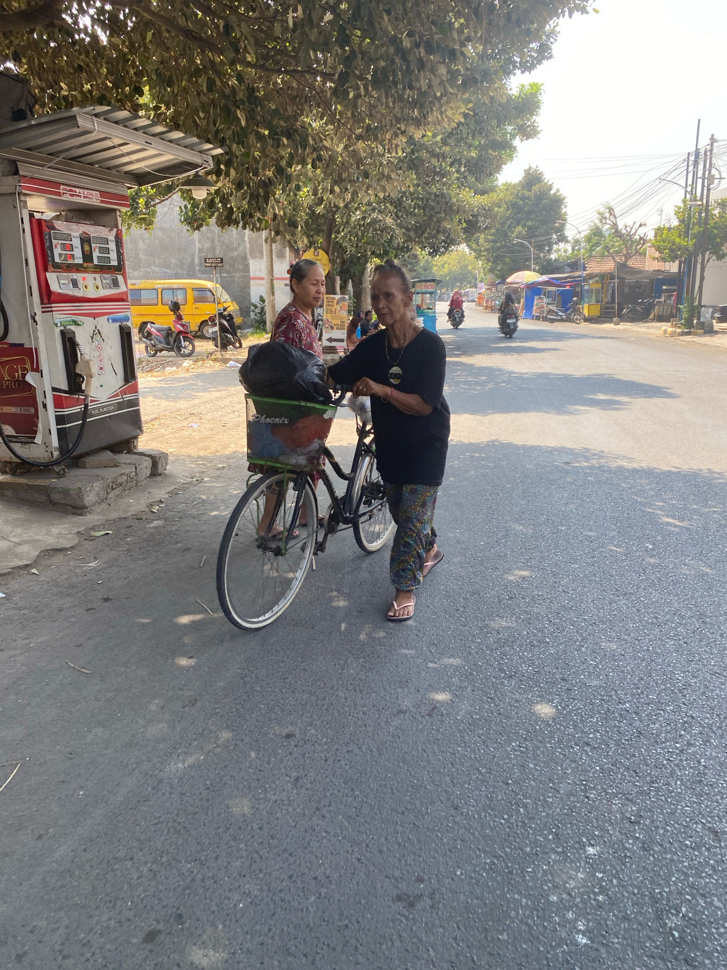 In Kediri City, Indonesia, a woman pushes a bicycle laden with a large black bag in its basket. She wears a dark t-shirt and patterned trousers, and her feet are adorned with pink flip-flops. She is walking on an asphalt road beside a row of roadside stalls and a gas station. Behind her, another woman, wearing a red and white patterned shirt, stands near the gas pump, looking away from the camera. In the background, a yellow van and several motorcycles are visible. The scene is set under a bright sky, with trees providing dappled shade.