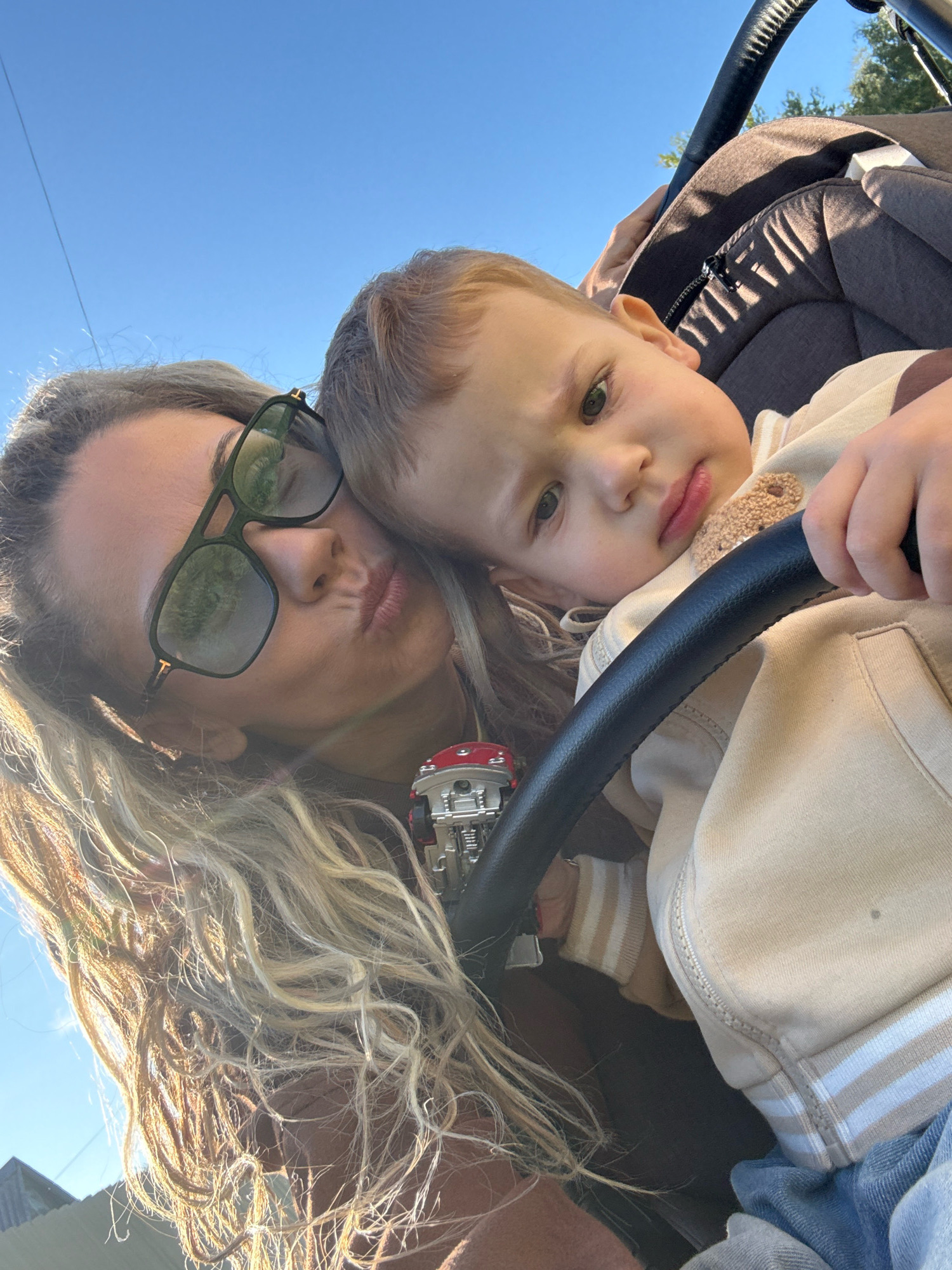A close-up selfie taken outdoors on a sunny day, featuring a woman with long blonde curly hair and sunglasses, making a kissing face towards the camera. She is leaning into a stroller where a young child with blonde hair is resting their head on her shoulder. The child is wearing a beige hooded jacket with striped cuffs. The background shows a clear blue sky and some green foliage. A stroller handle is prominently in the foreground.