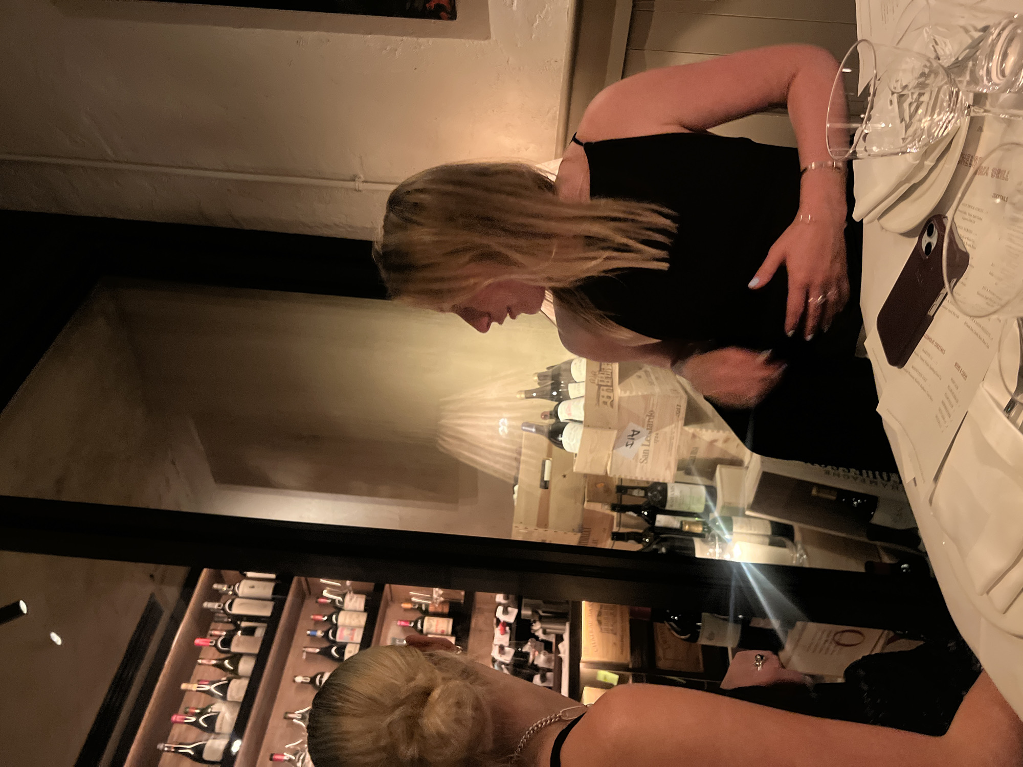 A brightly lit indoor restaurant scene at Chelsea Grill, London, features two women wearing black outfits seated at a table set with glasses, plates, a menu, and a phone. A glass wine display behind the table holds numerous bottles of wine and wooden boxes, reflecting the women's images.